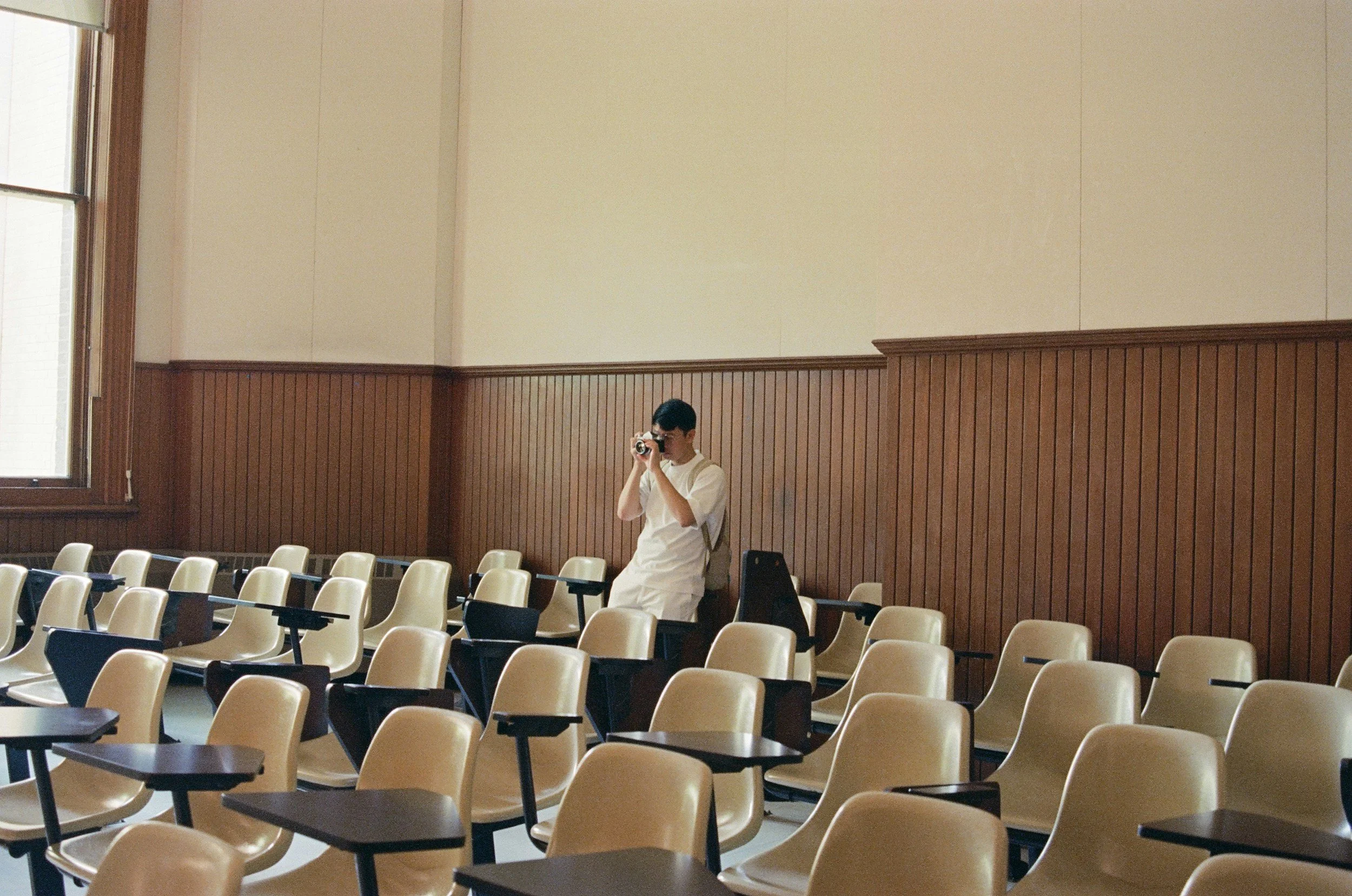 A person wearing white clothing taking a photo with a camera in an empty classroom or lecture hall with beige chairs and wooden paneling along the walls.