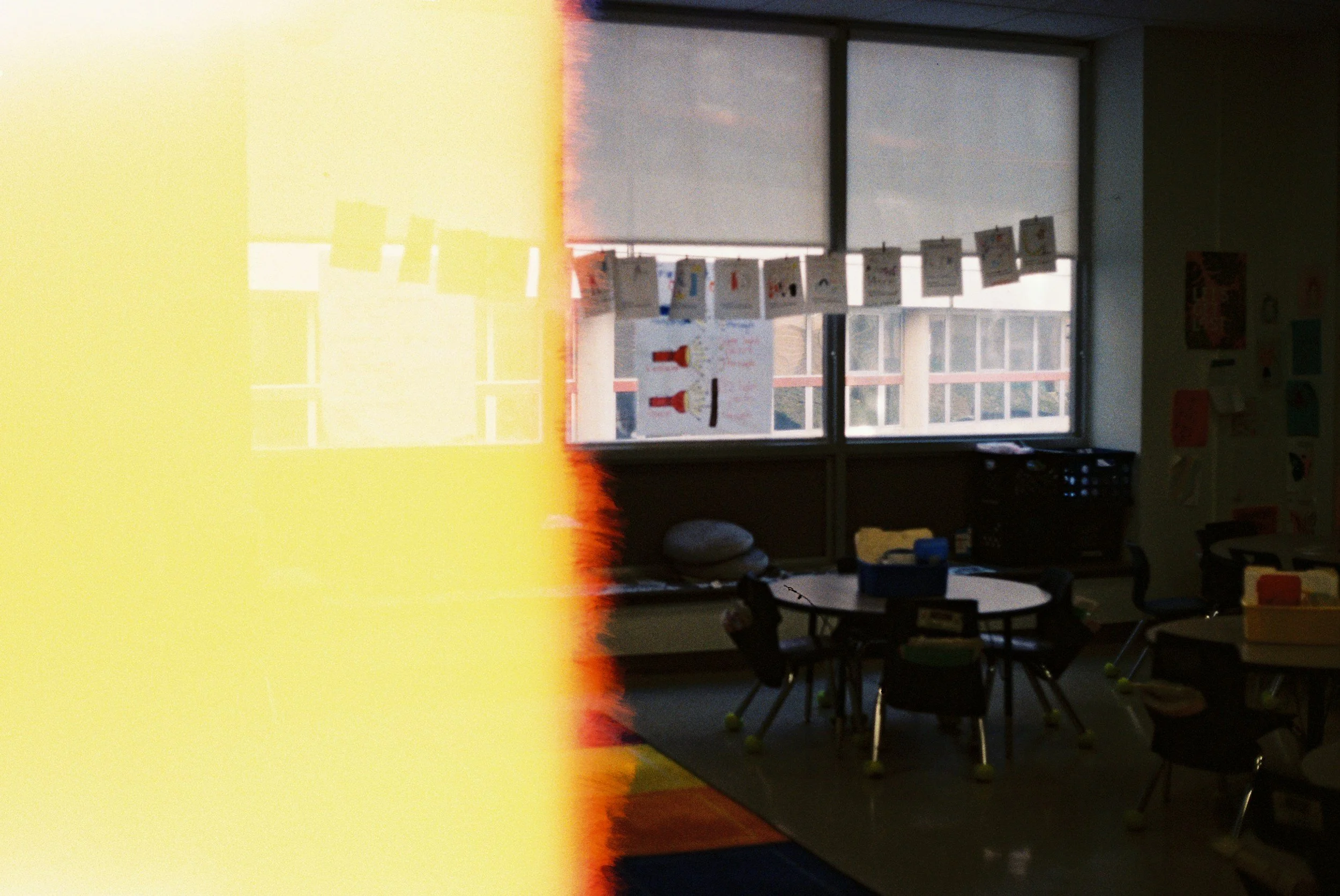 A classroom with tables and chairs, large windows, and student artwork hanging on a line. Part of the photo is obscured by a yellow and red light leak.