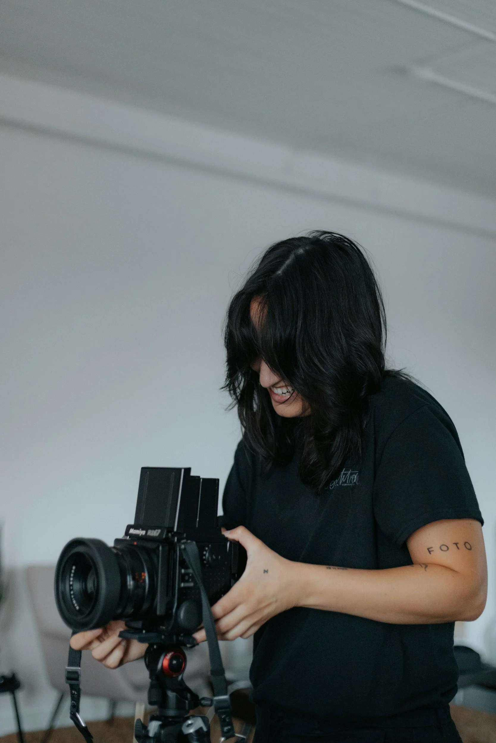 A woman with black hair and a tattoo on her left arm is smiling and adjusting a professional camera mounted on a tripod indoors.
