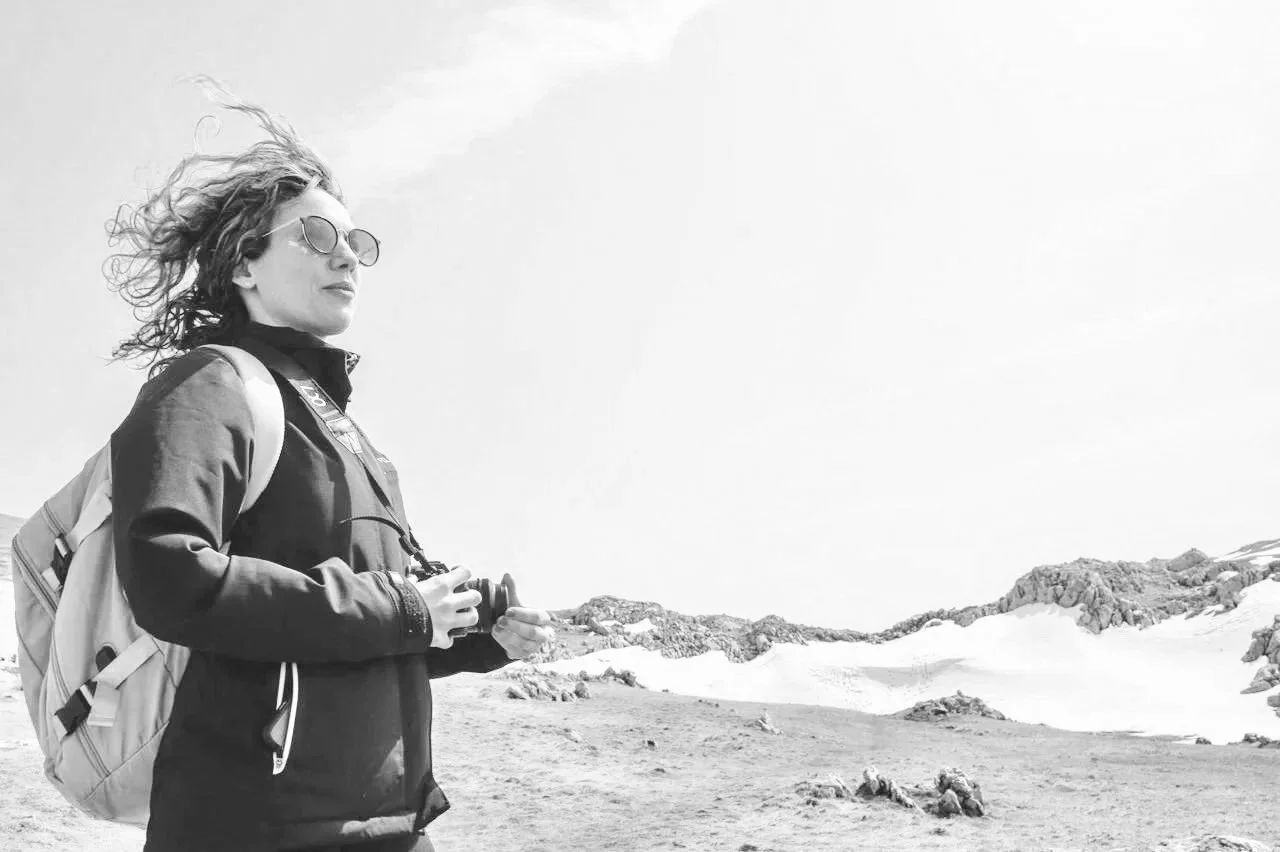 A woman with curly hair wearing sunglasses, a jacket, and a backpack holding binoculars, standing outdoors in a snowy mountain landscape.