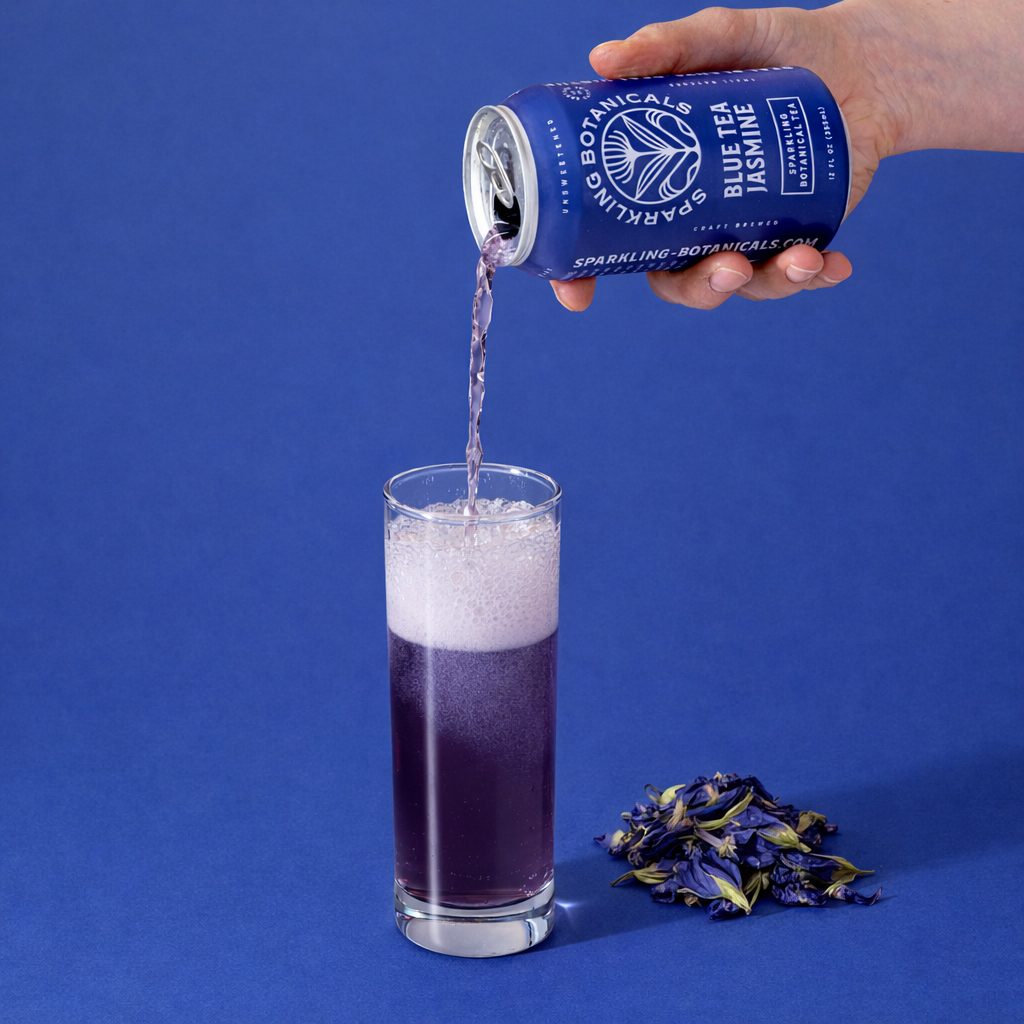 A person pouring sparkling purple soda from a can labeled 'Blue Botanical Jasmine' into a tall glass. There are dried purple and yellow flowers on the table in front of the glass, and a solid blue background.