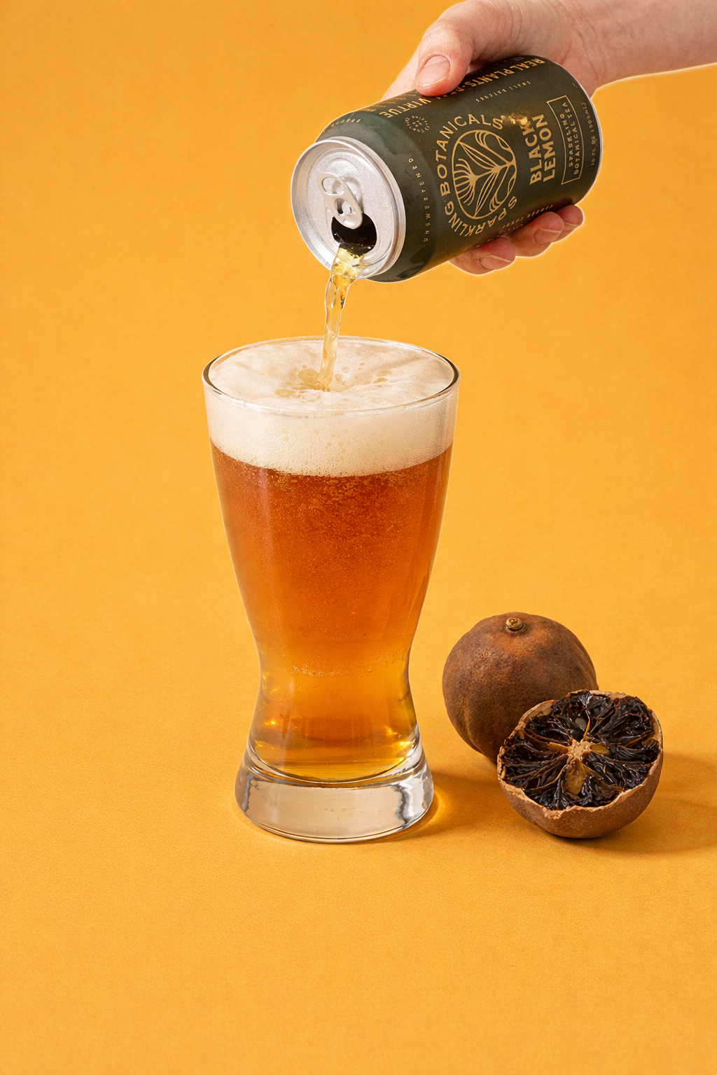 A hand pouring a can of botanical black lemon sparkling beverage into a glass, with a whole and halved black lemon on a yellow background.