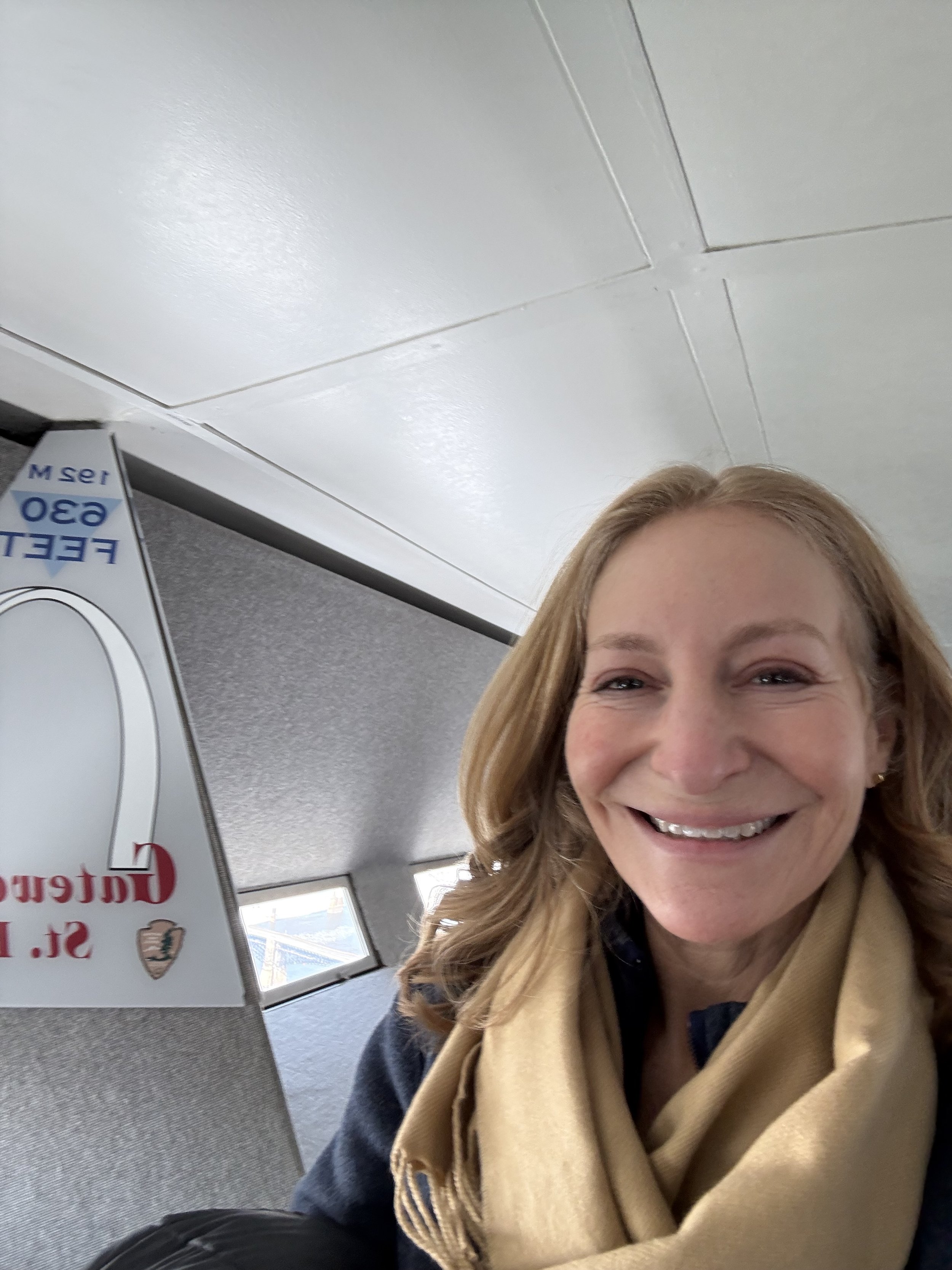 A smiling woman with red hair, glasses, and a beige sweater standing indoors in front of white cabinets.