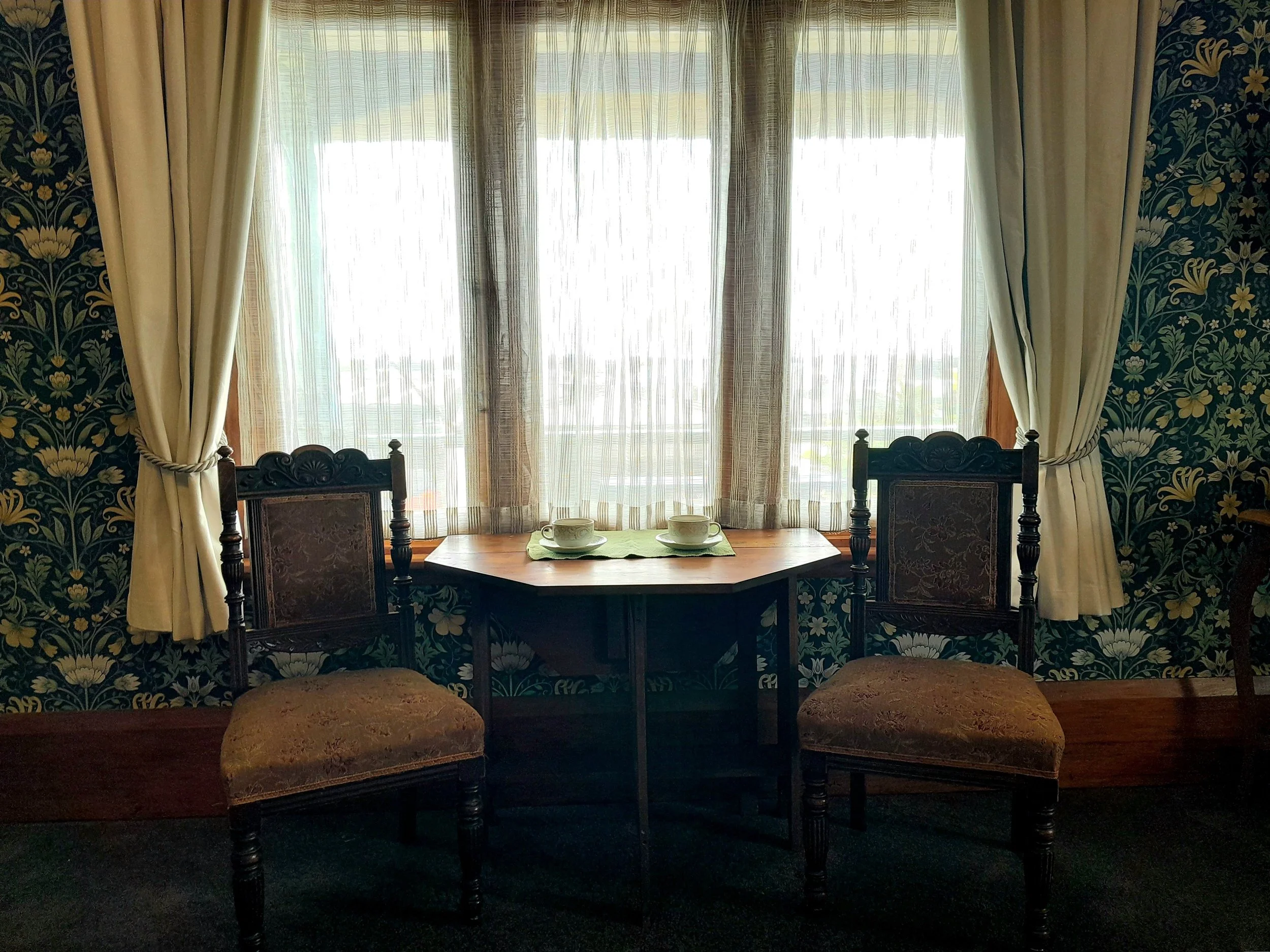 A cozy room with a window that has sheer curtains and cream-colored drapes, a small wooden table with two cups and green placemats, and two vintage chairs with dark wood frames and upholstered seats with floral patterns.