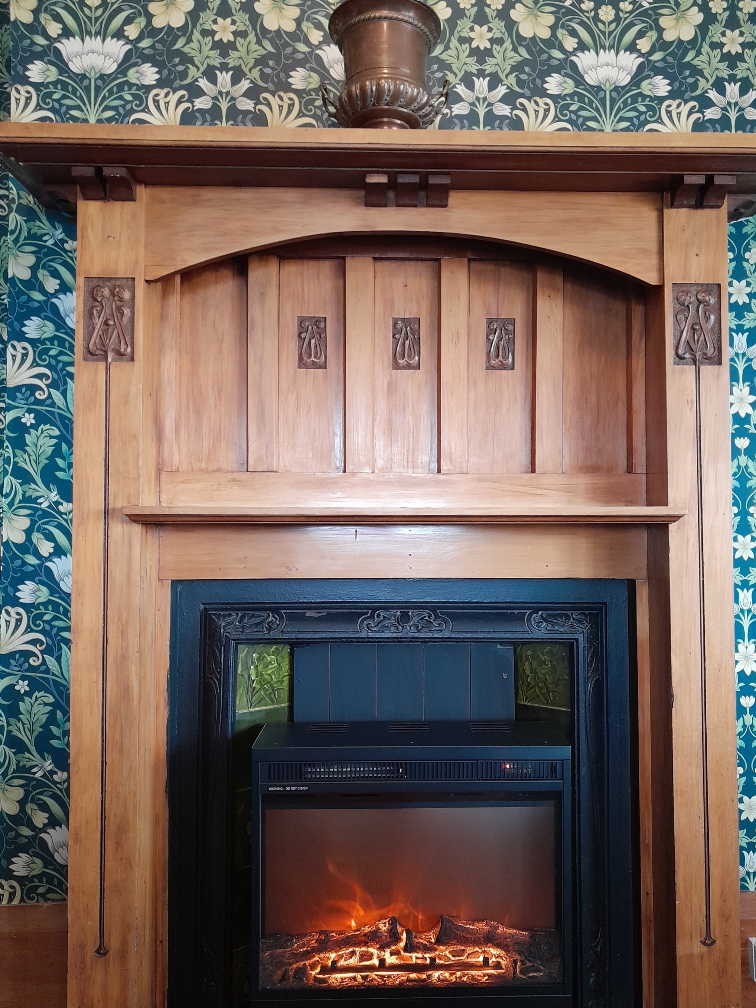 Wooden fireplace surround with decorative carvings and a black electric fireplace insert, set against floral wallpaper.