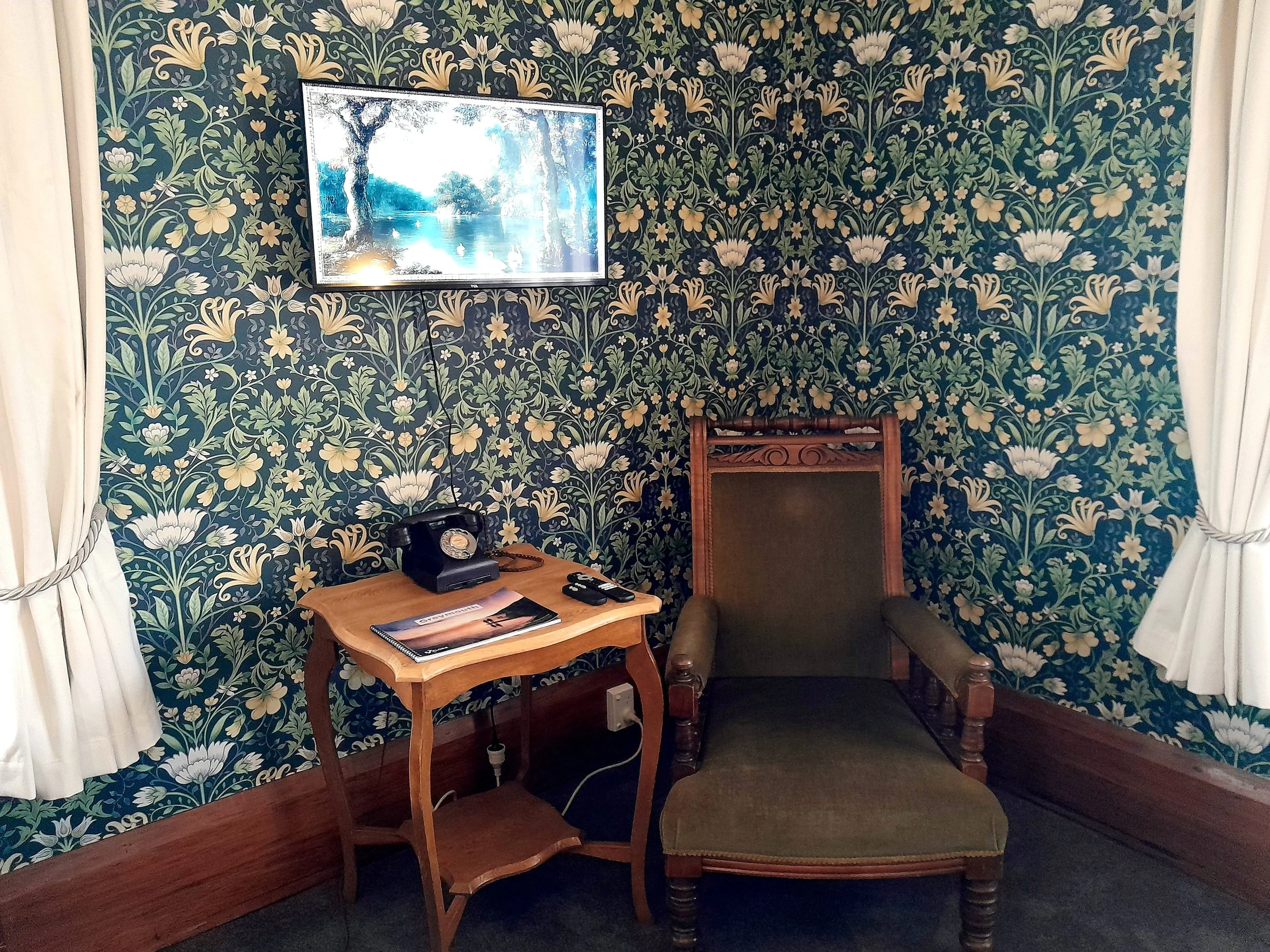 A cozy sitting area with floral wallpaper, an antique green armchair with wooden arms, a small wooden table holding a black rotary phone, a magazine, and a remote control. A flat-screen TV is mounted on the wall above the table, displaying a landscap