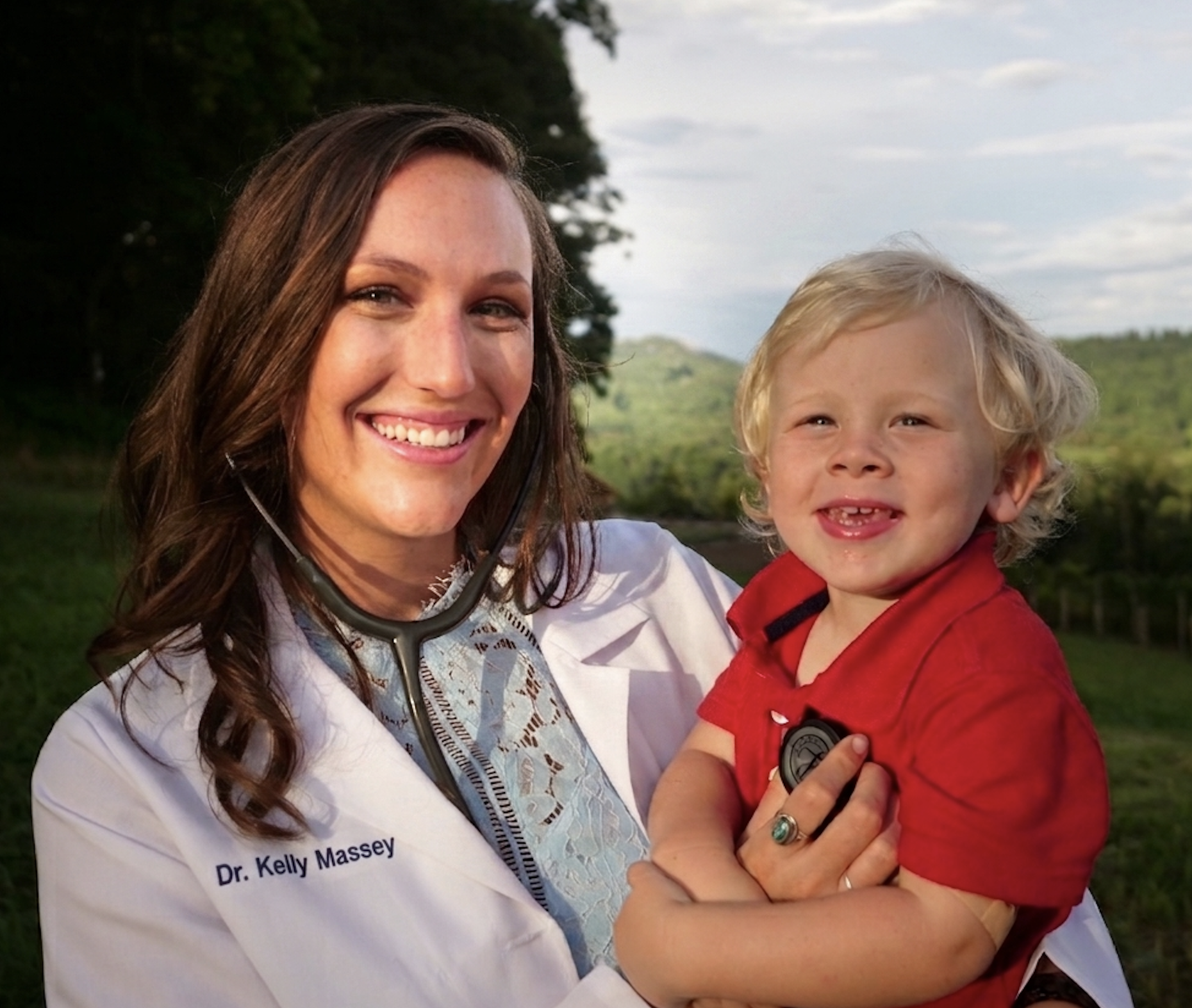 Dr. Kelly Massey naturopathic pediatric doctor in lab coat holding a smiling toddler with a stethoscope in the Palouse hills — natural pediatric care at Deeply Rooted Healing Haven Moscow Idaho