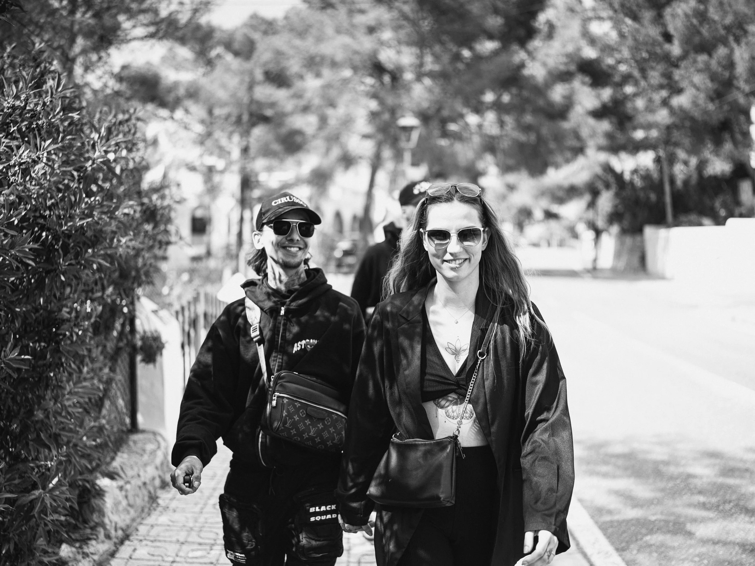 A couple walking outdoors, smiling, wearing sunglasses, with trees and a sidewalk in the background. during a family holiday Photographed by a professional dutch Photographer from Eindhoven