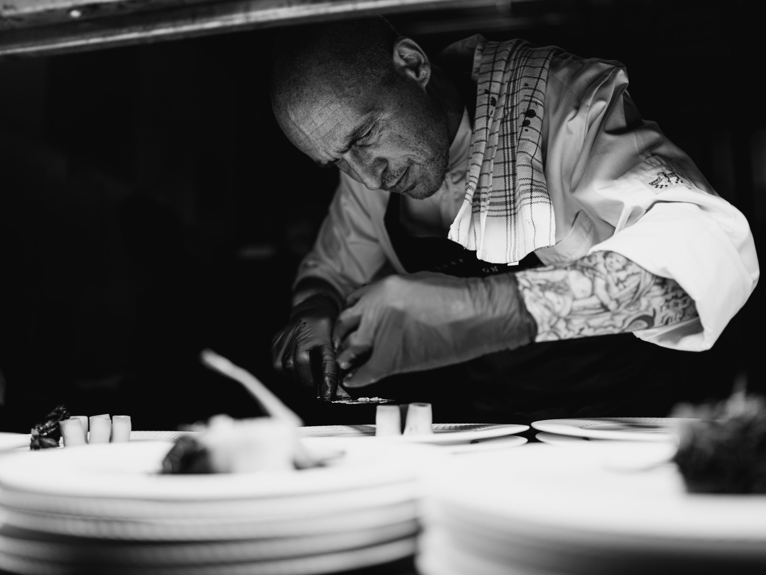 Black and white photo of a chef in a professional kitchen, hunched over and meticulously plating a dish on a plate, with similar plates stacked in the foreground.