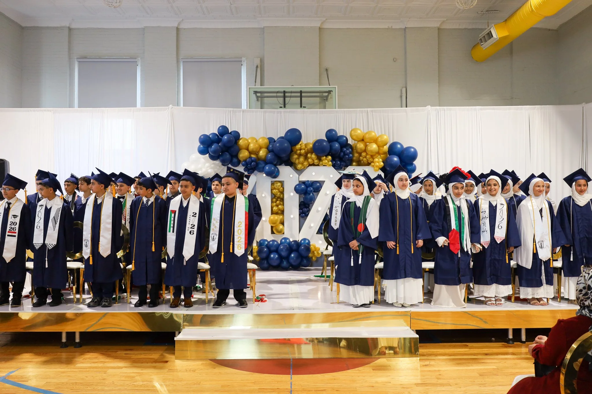 Group of students in graduation caps and gowns standing on a stage decorated with blue, gold, and white balloons at a graduation ceremony.