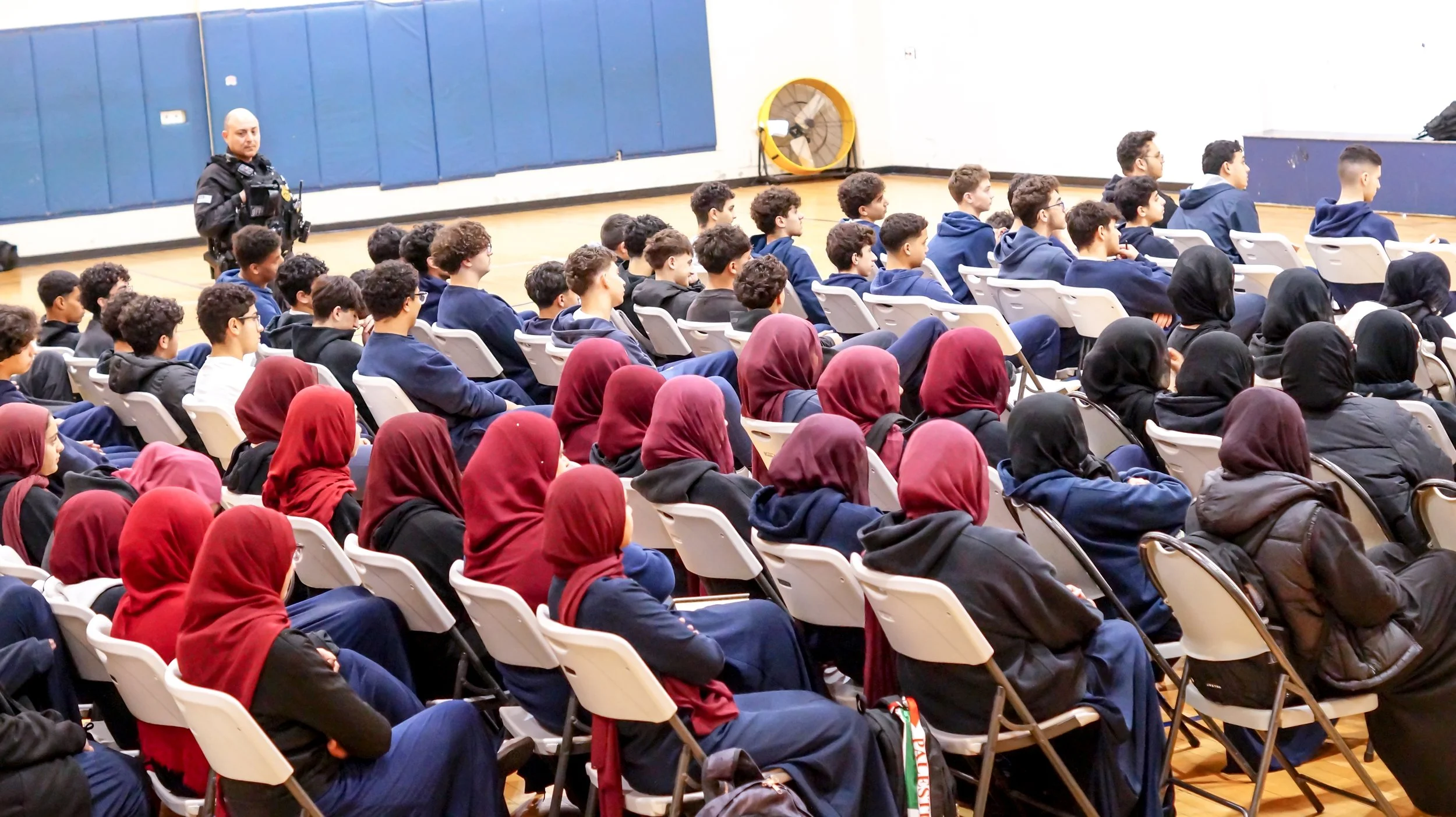 A group of students seated in a gymnasium, listening to a police officer during what appears to be an educational presentation.