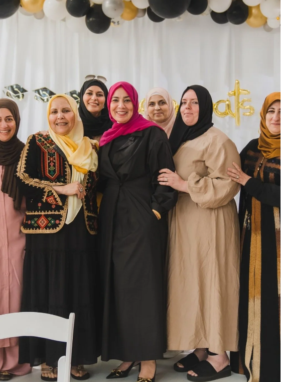 Group of women dressed in colorful outfits and hijabs at celebratory event with balloons and decorations in the background.