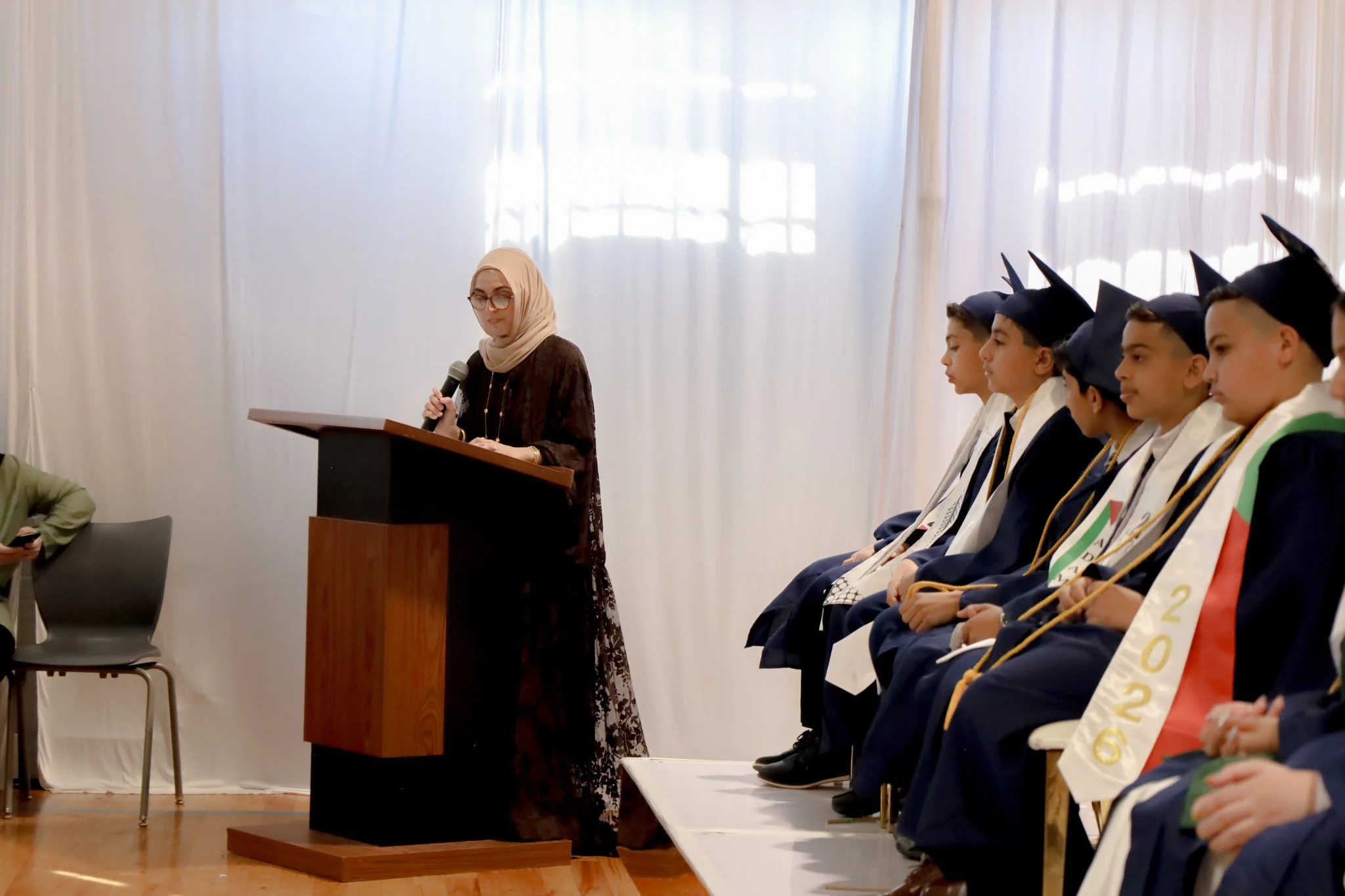 A woman in a beige hijab and glasses stands at a podium, speaking into a microphone during a graduation ceremony. She is facing a line of young male graduates wearing caps and gowns, seated in chairs, with some wearing sashes and medals. The background features white curtains and bright natural light.