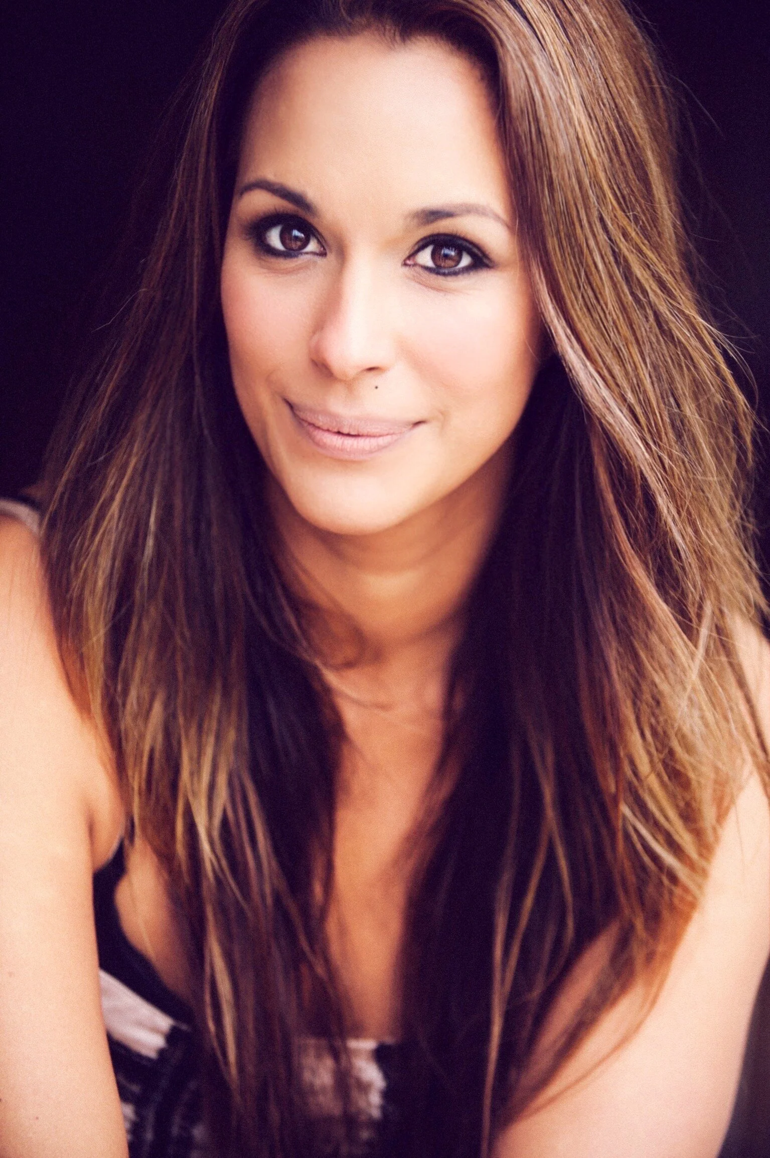 A woman with long brown hair, wearing makeup, smiling slightly, against a dark background.