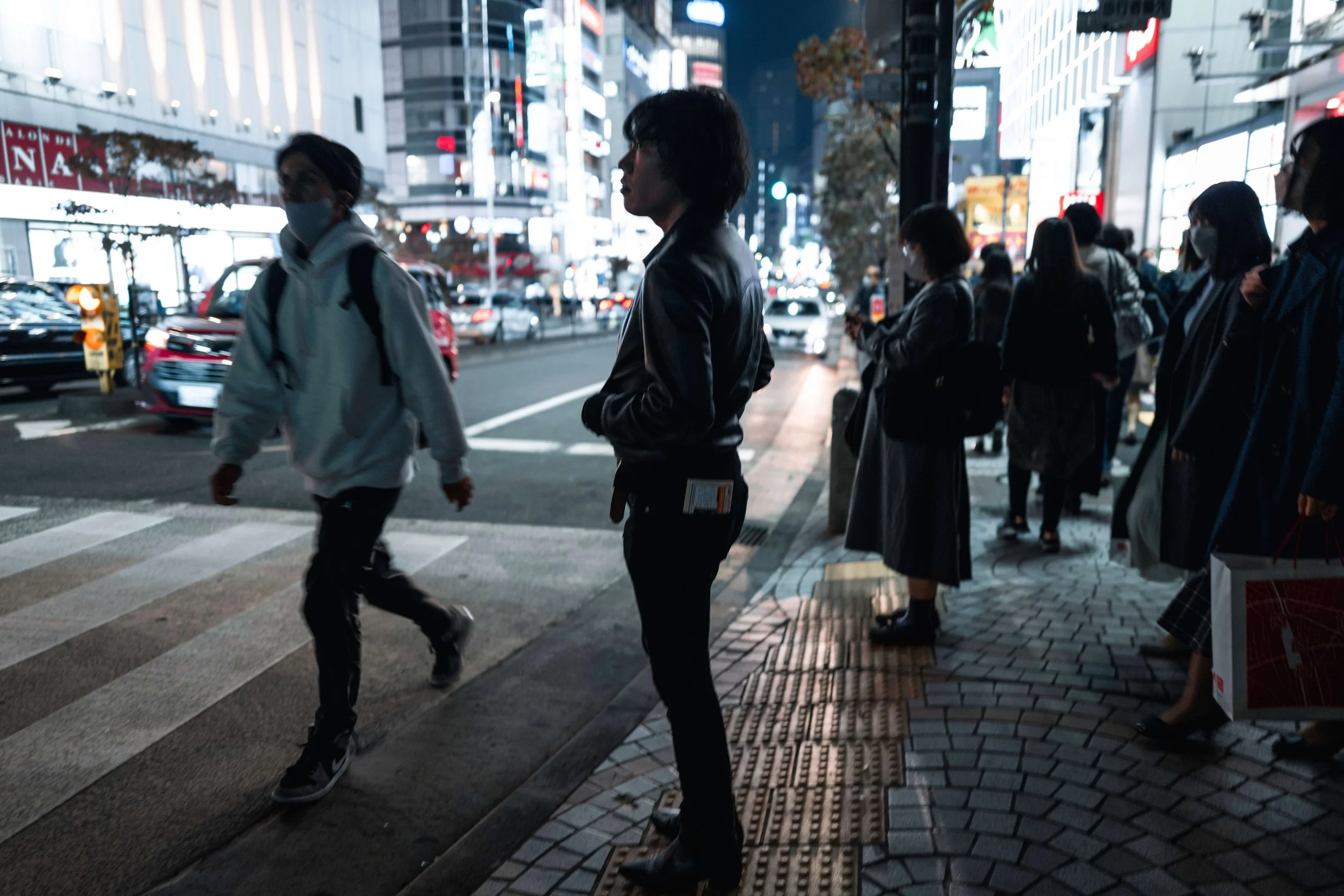 People waiting at a crosswalk on a busy city street at night, with illuminated buildings and cars passing by.
