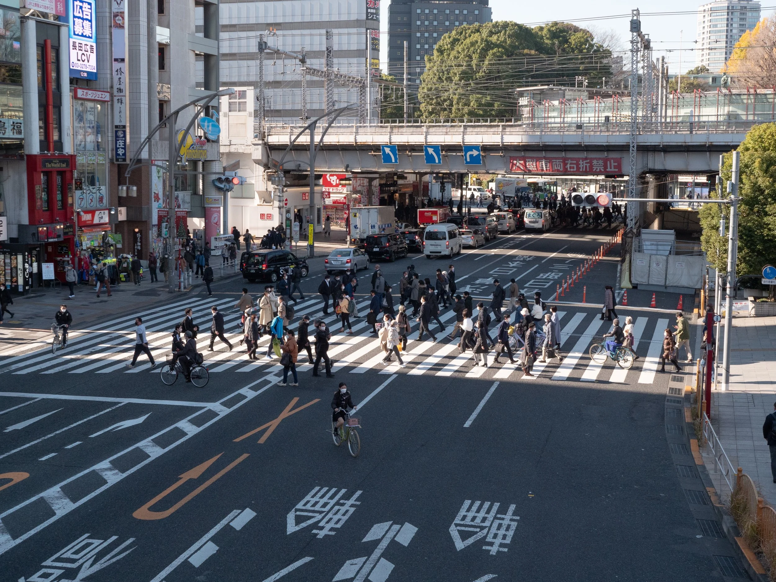 A busy crosswalk in an urban city, with pedestrians and cyclists crossing the street, high-rise buildings, and an overpass above. Cars are waiting at traffic signals, and storefronts line the street.