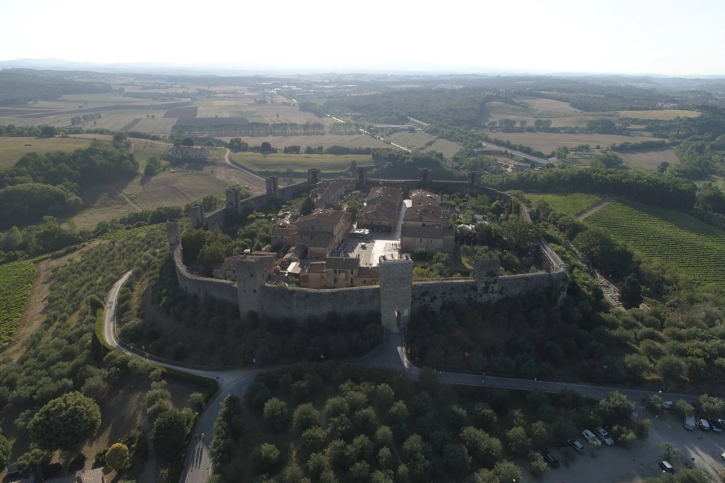 Aerial view of a historic hilltop castle with defensive walls and towers, surrounded by lush green countryside in Italy.