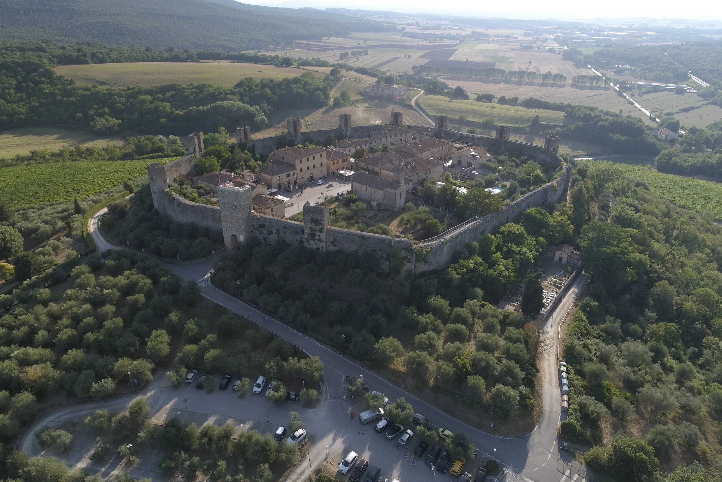 Aerial view of an ancient walled fortress with towers, surrounded by lush green landscape, roads, and farmland.