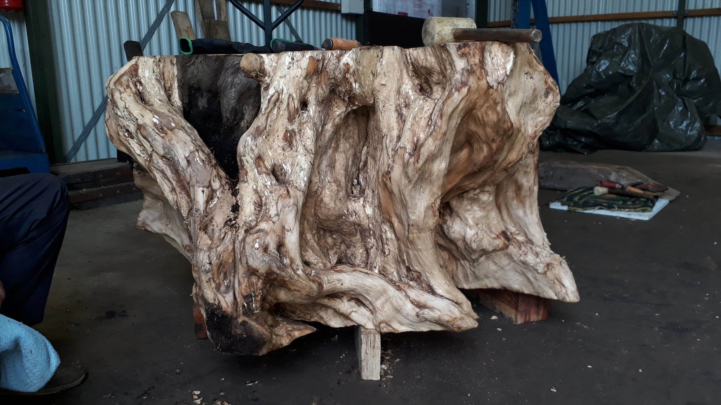 A large piece of natural, textured wood with many knots and crevices, resting on small supports inside a workshop with tools and equipment in the background.