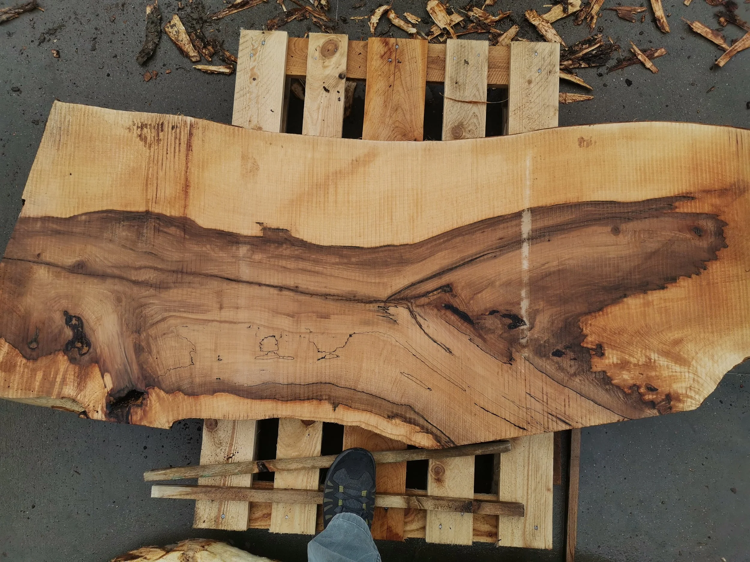 A wooden table top with natural edges, placed on a pallet for support, surrounded by wood shavings and small pieces of wood on a concrete surface.