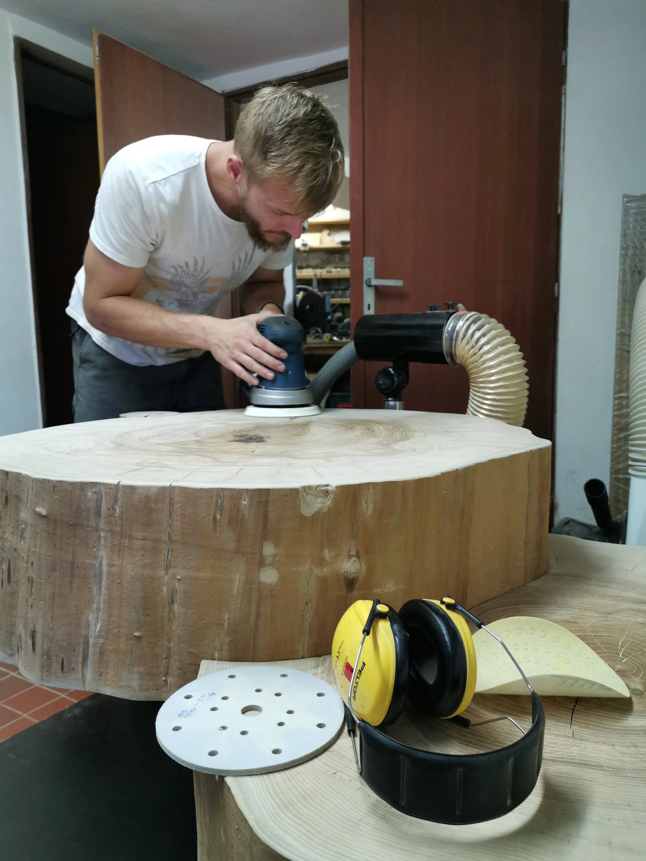 A man is using a handheld sander on a large piece of wood in a woodworking workshop. The workspace includes various tools and equipment, with protective ear muffs placed on the table.