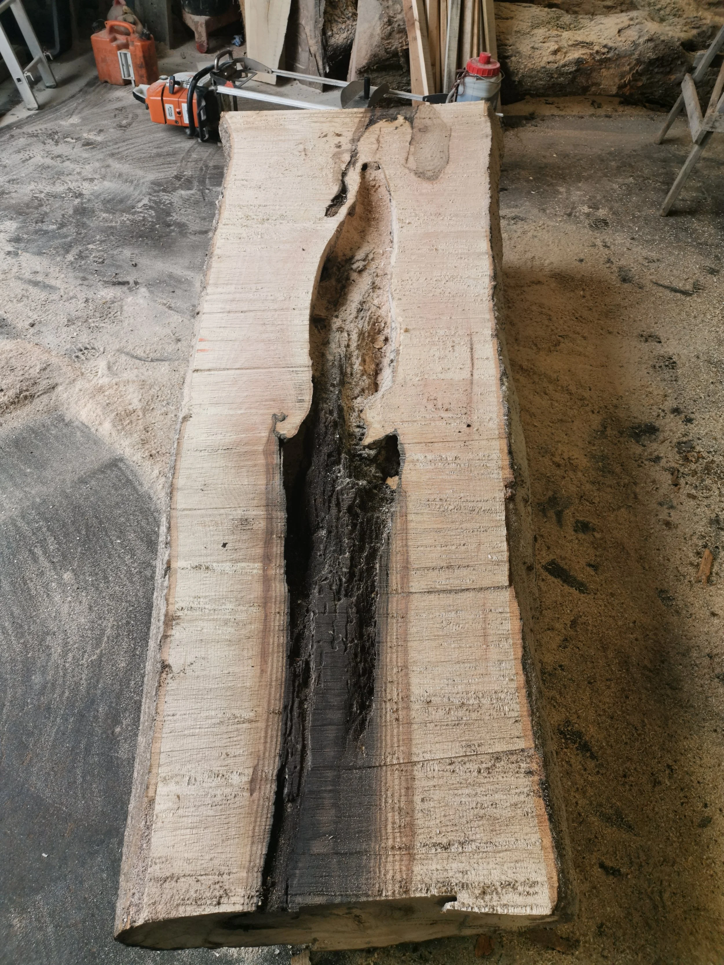 A large wooden log with a natural split, placed in a workshop. Equipment such as a chainsaw and other tools are visible in the background.