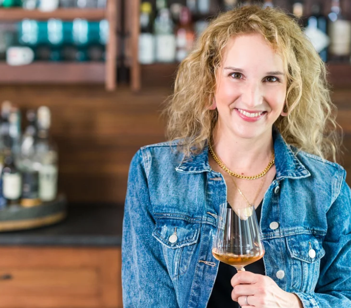 A woman with curly blonde hair smiling and holding a glass of whiskey in a bar or tasting room