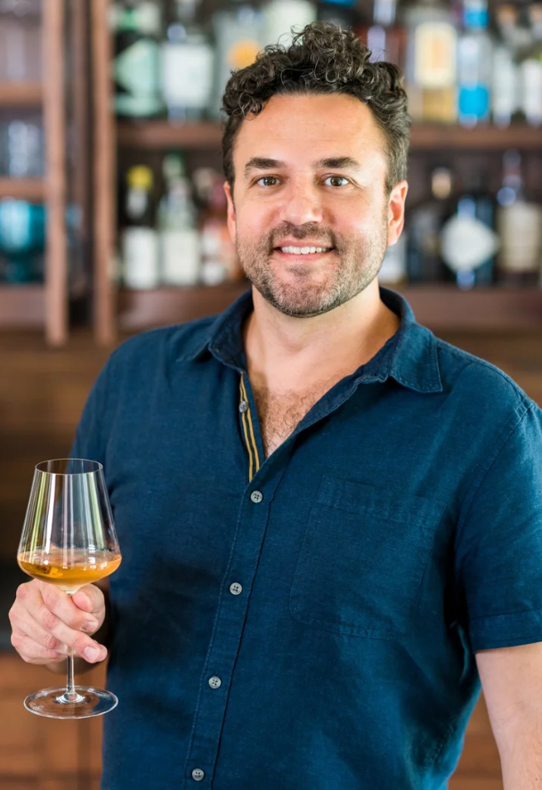Man with curly dark hair and a beard holding a glass of white wine, standing in front of a wooden bar with bottles of alcohol.