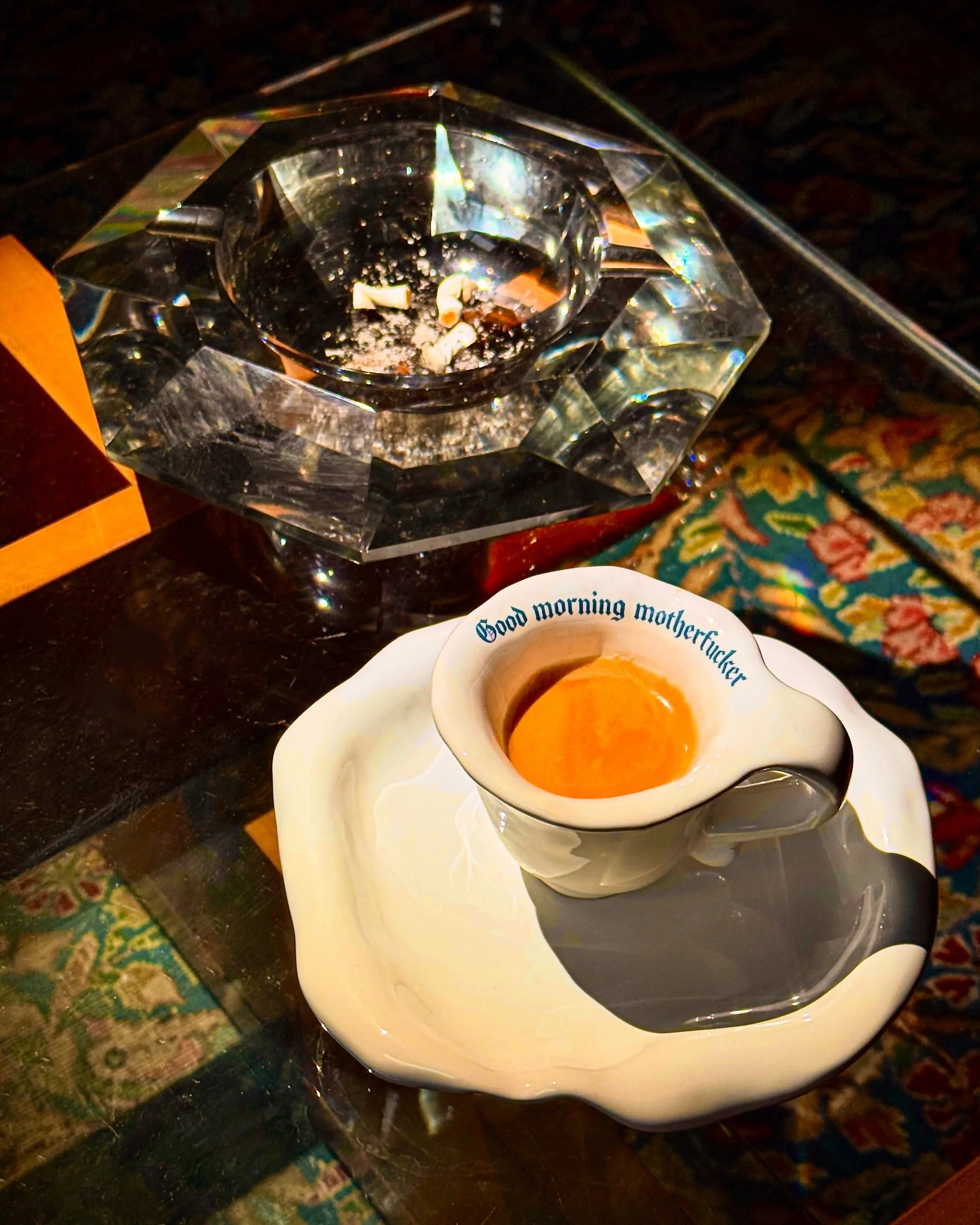 A coffee cup with the inscription 'Good morning motherfucker' on the rim, filled with coffee, placed on a floral dish. Behind it, there is a glass ashtray with cigarette ashes and remnants, atop a dark wooden surface with a colorful patterned cloth n