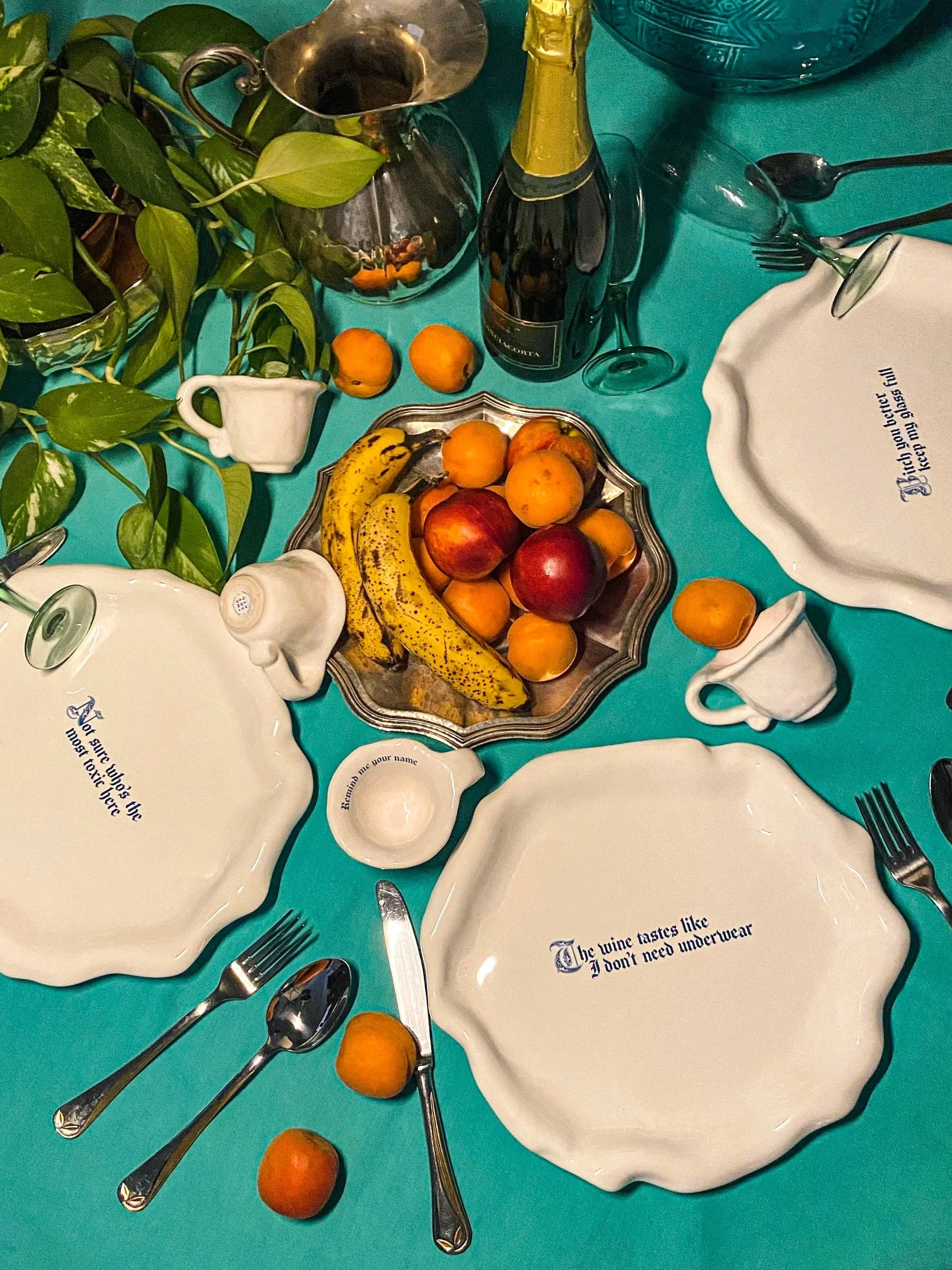 A dining table set with plates, cutlery, and cups on a turquoise tablecloth. The plates have printed sayings, including 'The wine tastes like I don't need underwear.' A large silver tray at the center holds bananas, apples, peaches, and plums. There 