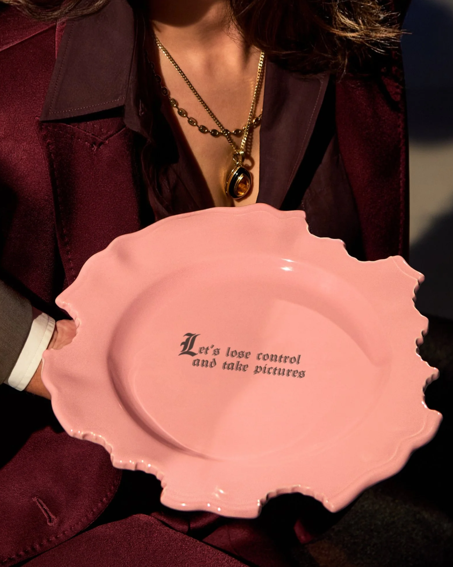 A woman with styled brown hair, wearing a burgundy blazer, holding a pink scalloped plate with the text 'Let's lose control and take pictures' written on it.