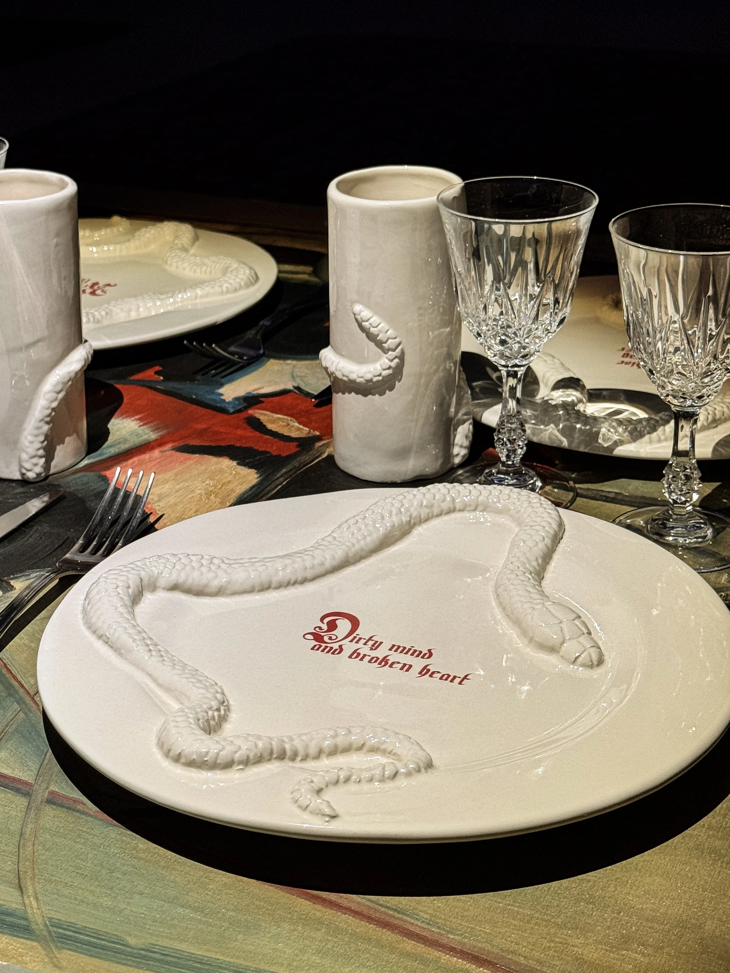 A white plate with a snake-shaped decoration and the phrase 'Dirty mind and broken heart' written on it, two crystal glasses, white candles in ceramic holders, and a multicolored tablecloth.
