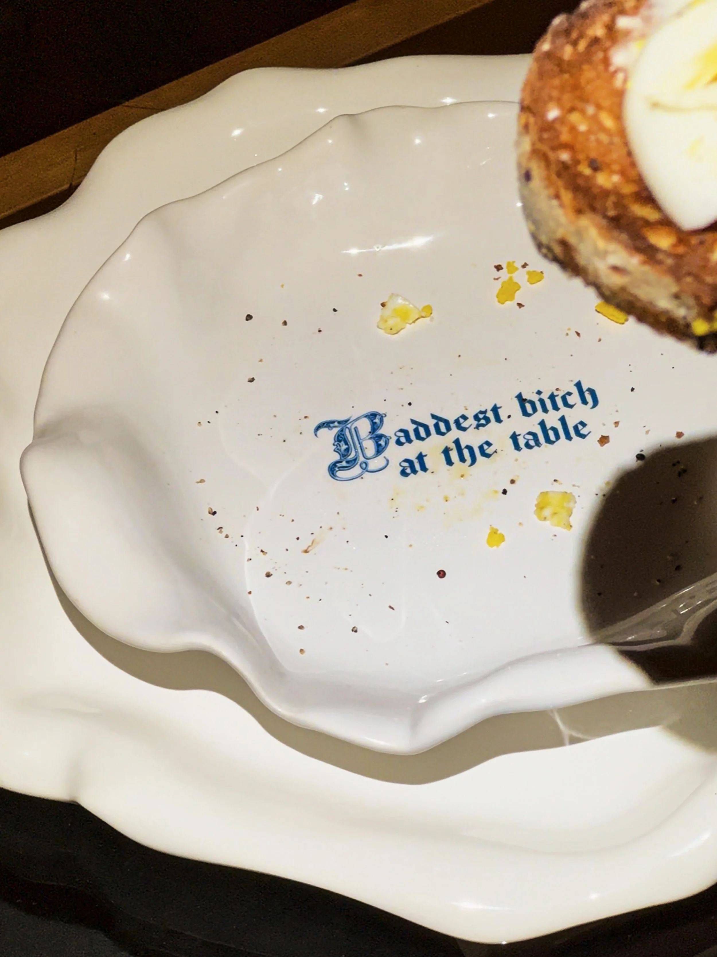 Empty white dish with the phrase 'Baddest bitch at the table' in blue text, crumbs and bits of food inside, partially obscured by a spoon and a piece of food.