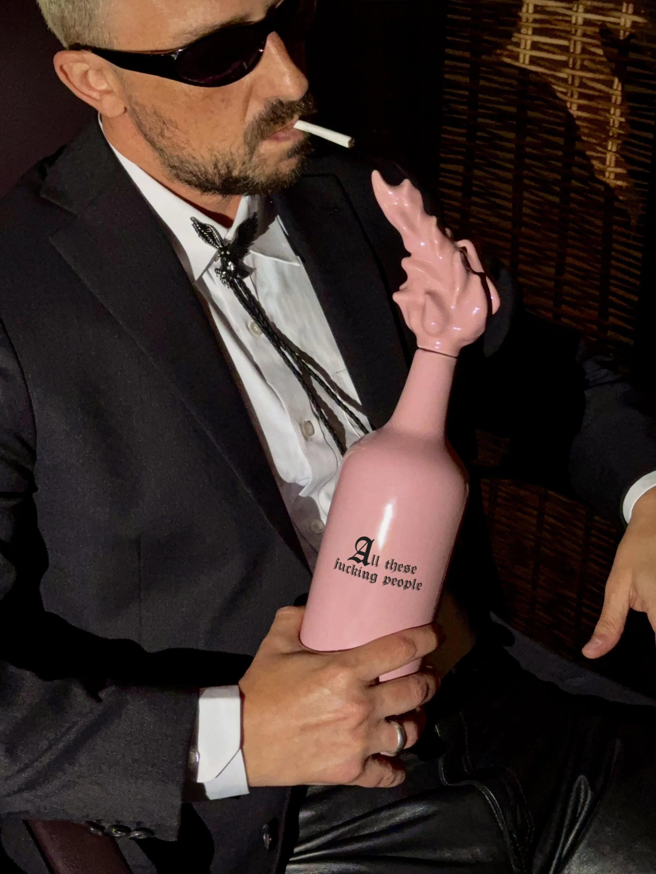 A man in a tuxedo with sunglasses is smoking a cigarette and holding a pink dildo with the text 'All these fucking people' written on it.