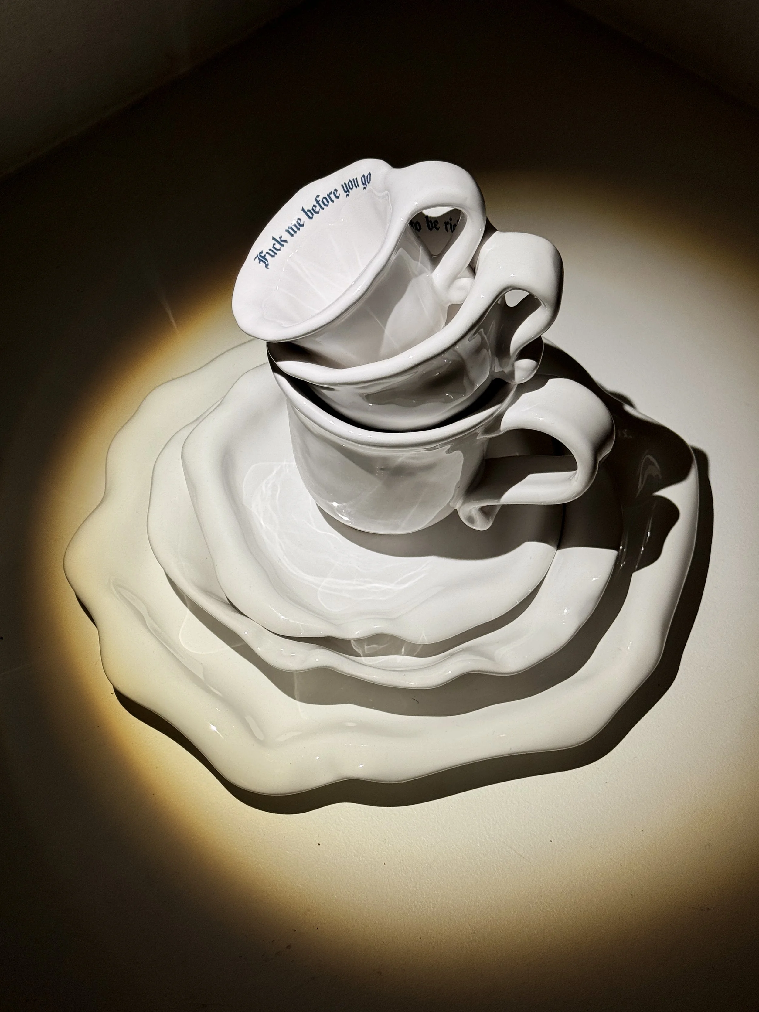Stacked white porcelain teacups and saucers with a quote on the top cup that reads, 'F*** me before you go to be r***.'
