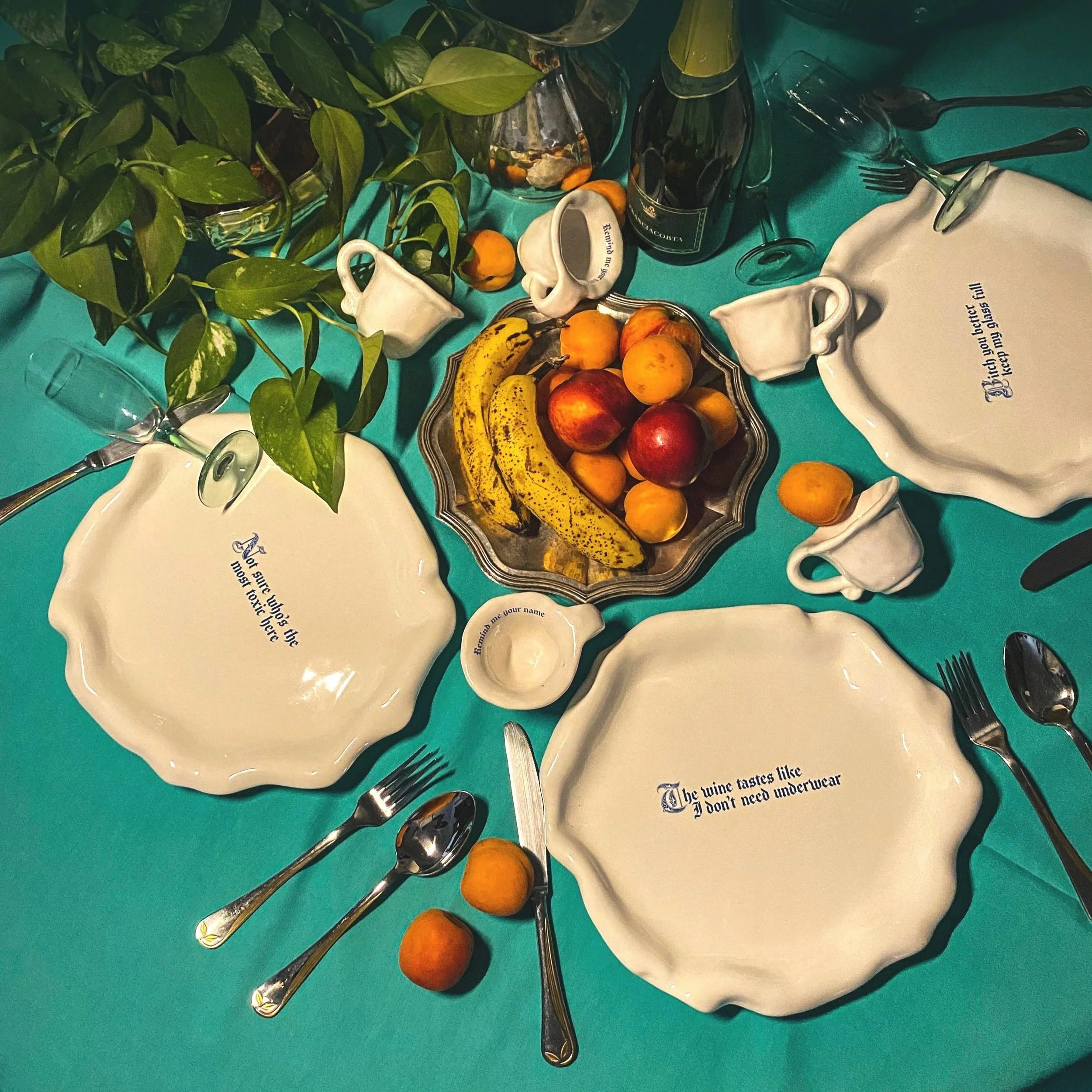 A dining table set with white ceramic plates, silverware, and glasses. There are green leaves and a variety of fruits including bananas, apples, and peaches on the table. The tablecloth is teal, and there are decorative text plates with quotes about wine and clothing.