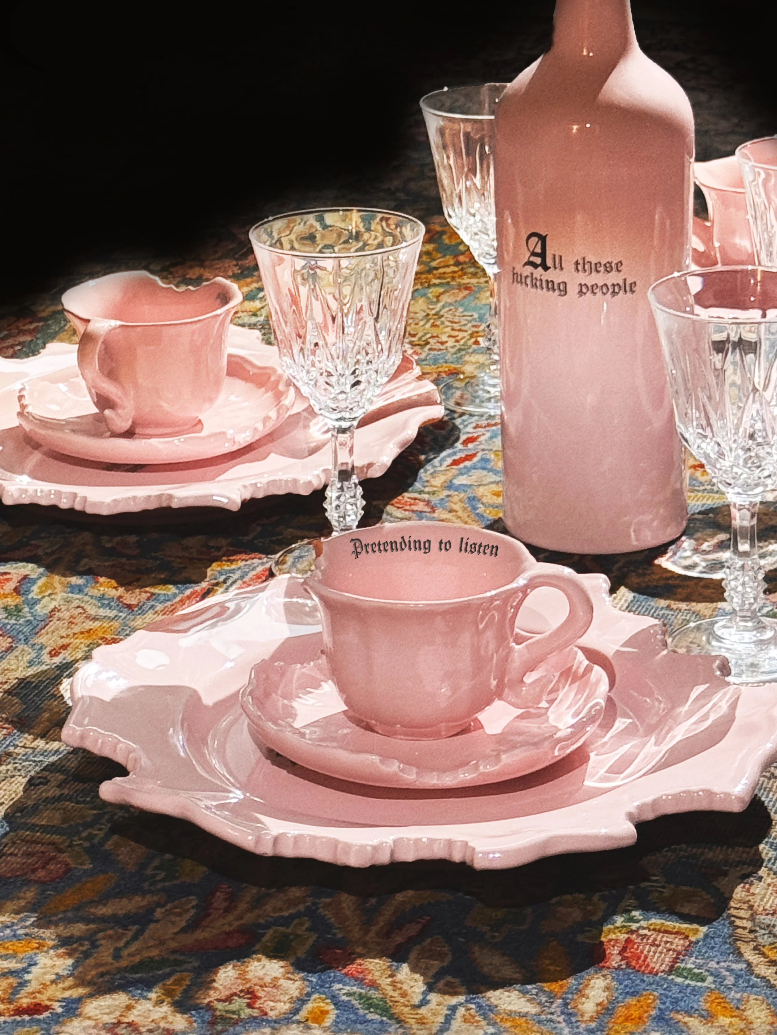 A pink ceramic teacup and saucer on a pink ceramic plate, set on a decorative cloth, with a pink bottle and clear glasses in the background.