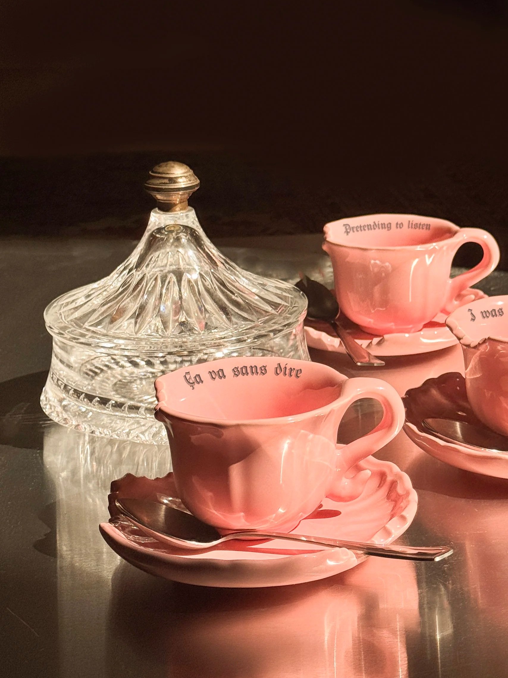 Pink teacups and saucers with inscribed phrases, glass sugar bowl, and a spoon on a reflective surface.