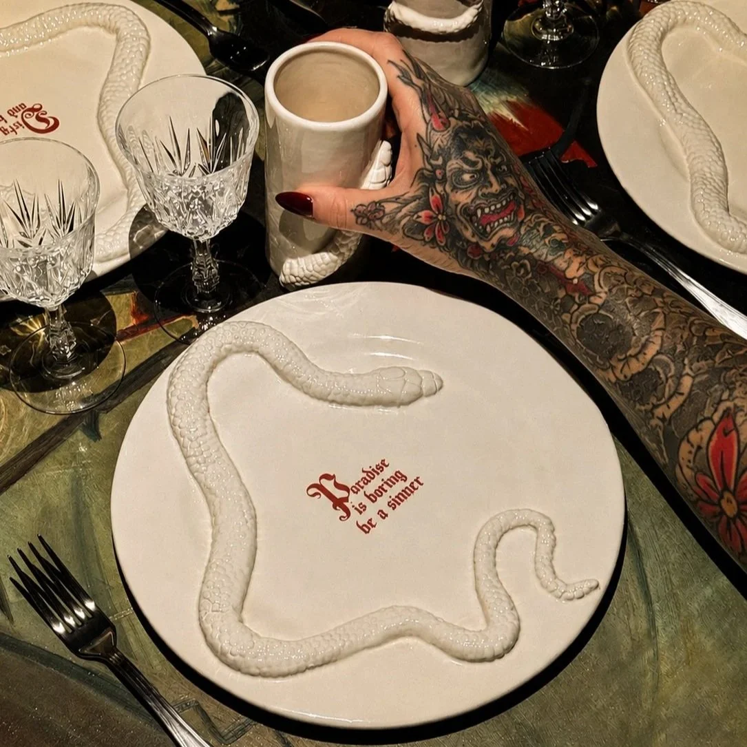 A dinner table setting with a white plate featuring a snake-shaped design made of a white substance, and red text reading 'Paradise is boring for a sinner' in the center. There are two crystal wine glasses, a beige mug, and a hand with colorful tattoos grasping the mug. The table has additional plates, cutlery, and decorative items.
