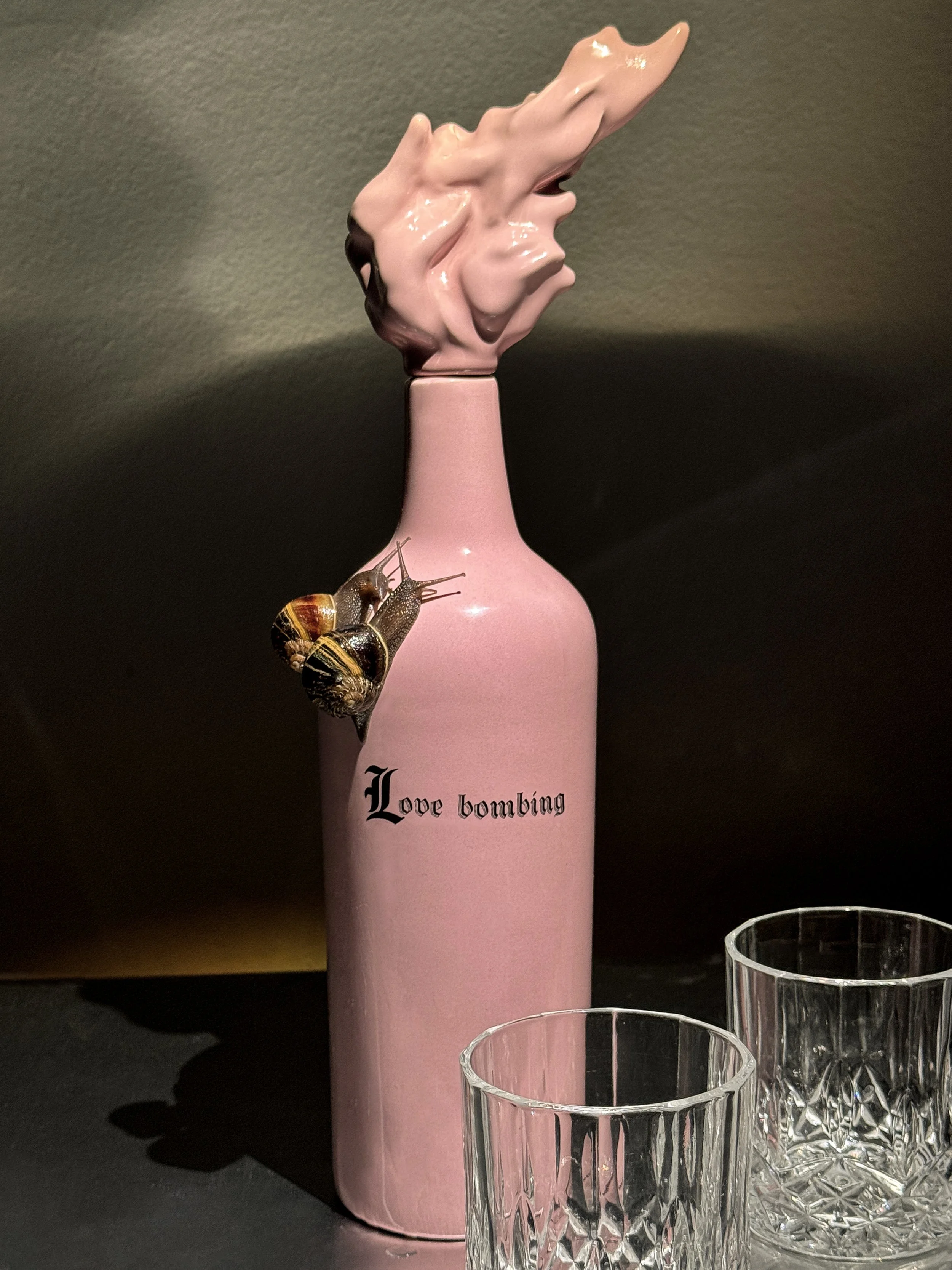 A pink bottle with the words 'Love bombing' written on it, a couple of snails on it, and two empty clear glass tumblers on a dark surface.