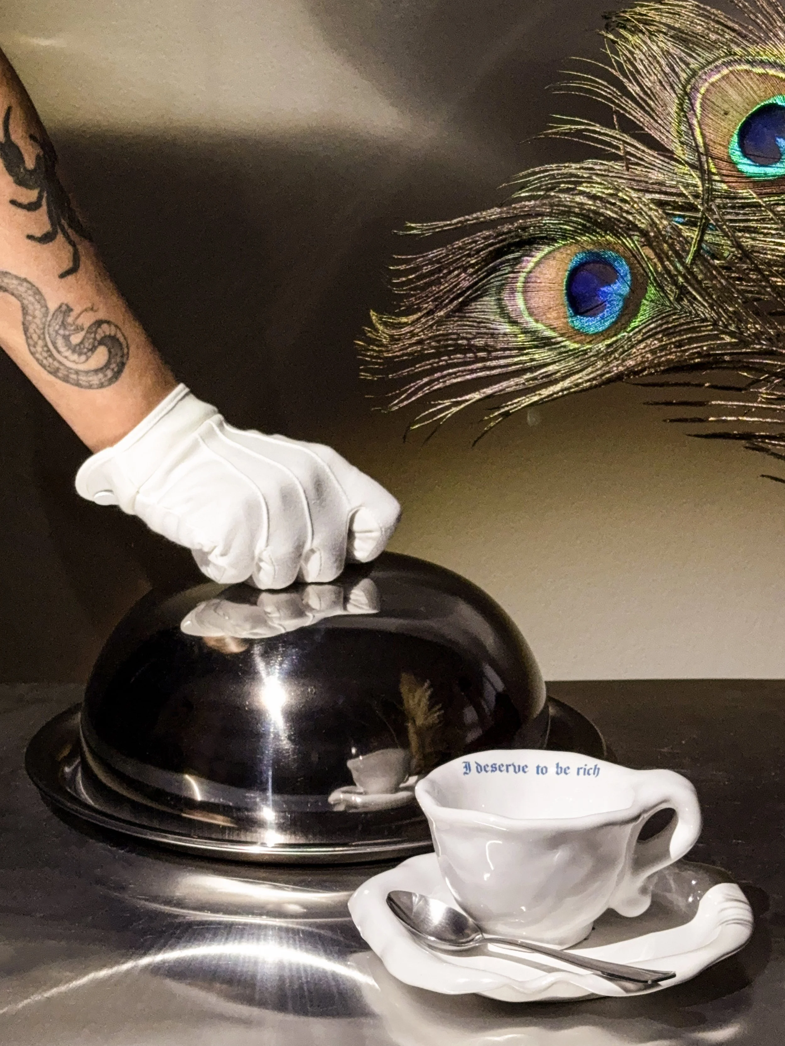 A person wearing a white glove is lifting a silver lid on a serving tray. A decorative peacock feather and a porcelain cup with a spoon, inscribed with the words "I deserve to be rich," are also visible.