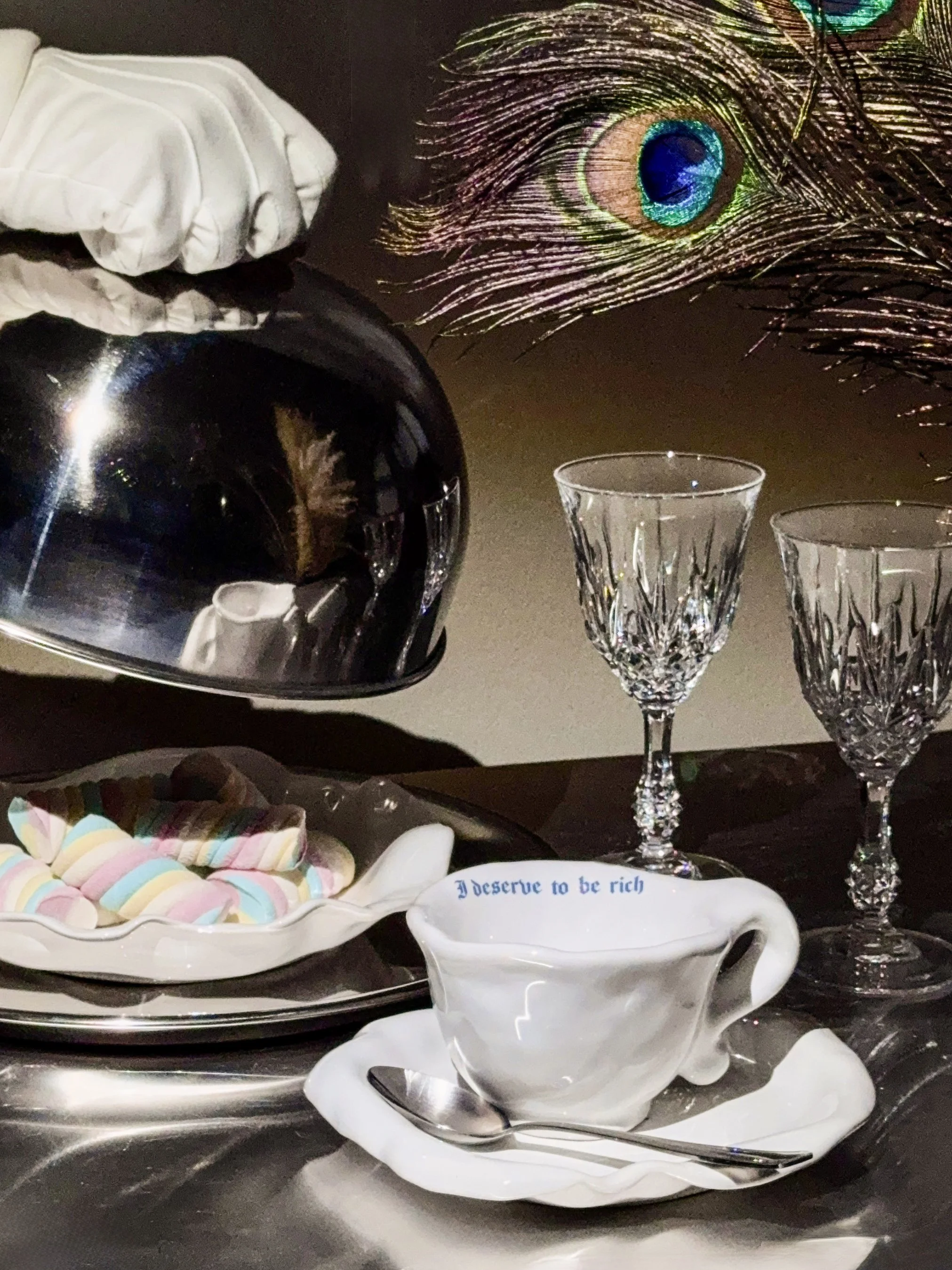 A table setting with a teapot, two crystal glasses, a white dish with pastel-colored marshmallows, and a white teacup with blue writing that reads 'I deserve to be rich.' A peacock feather and a shiny black helmet are also on the table.