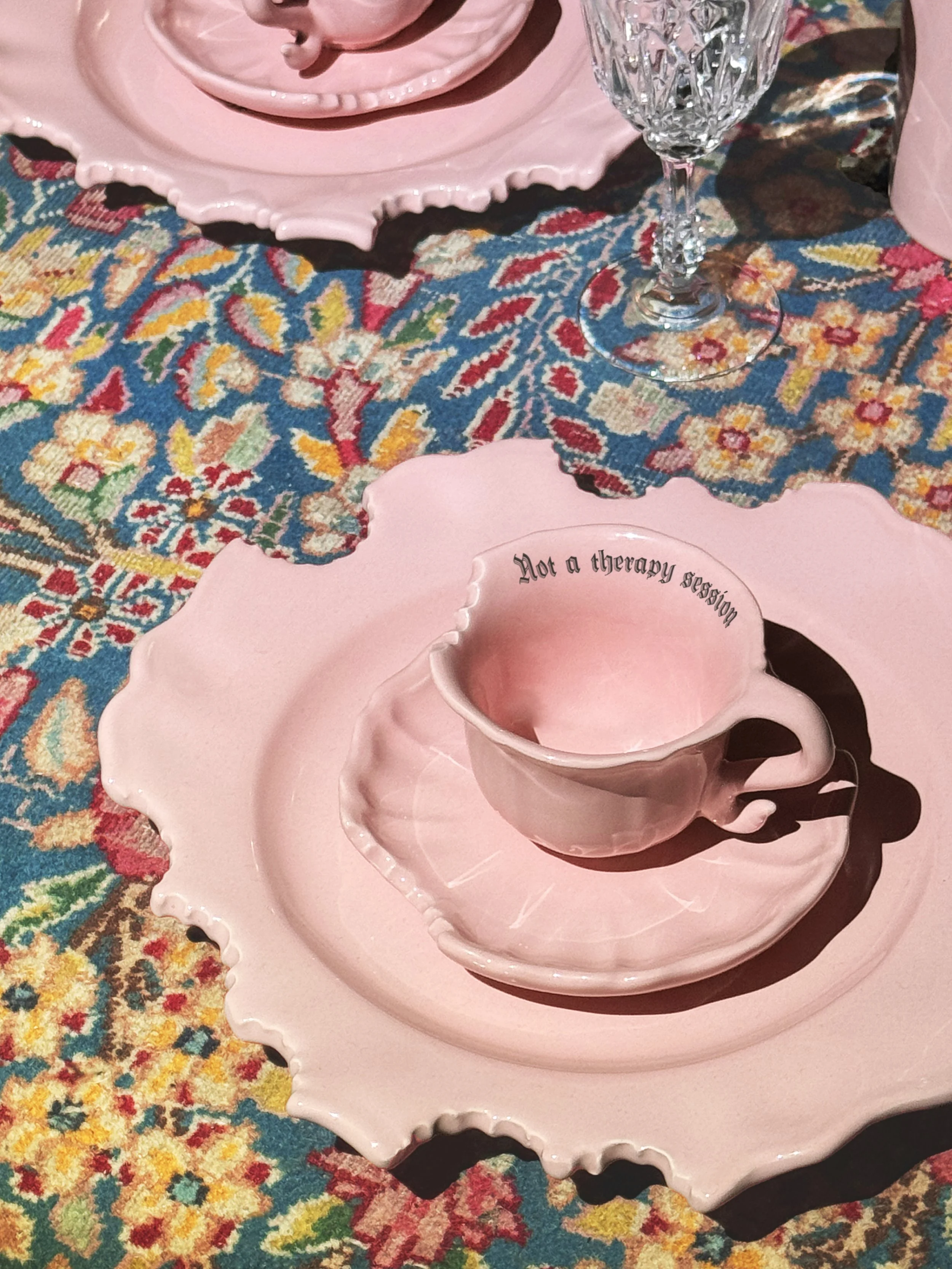 Pink ceramic teacup with a quote, resting on a matching pink saucer, placed on a decorative pink tray. The tray is on a colorful, floral-patterned fabric surface. The quote inside the cup reads, 'Not a therapy session.'