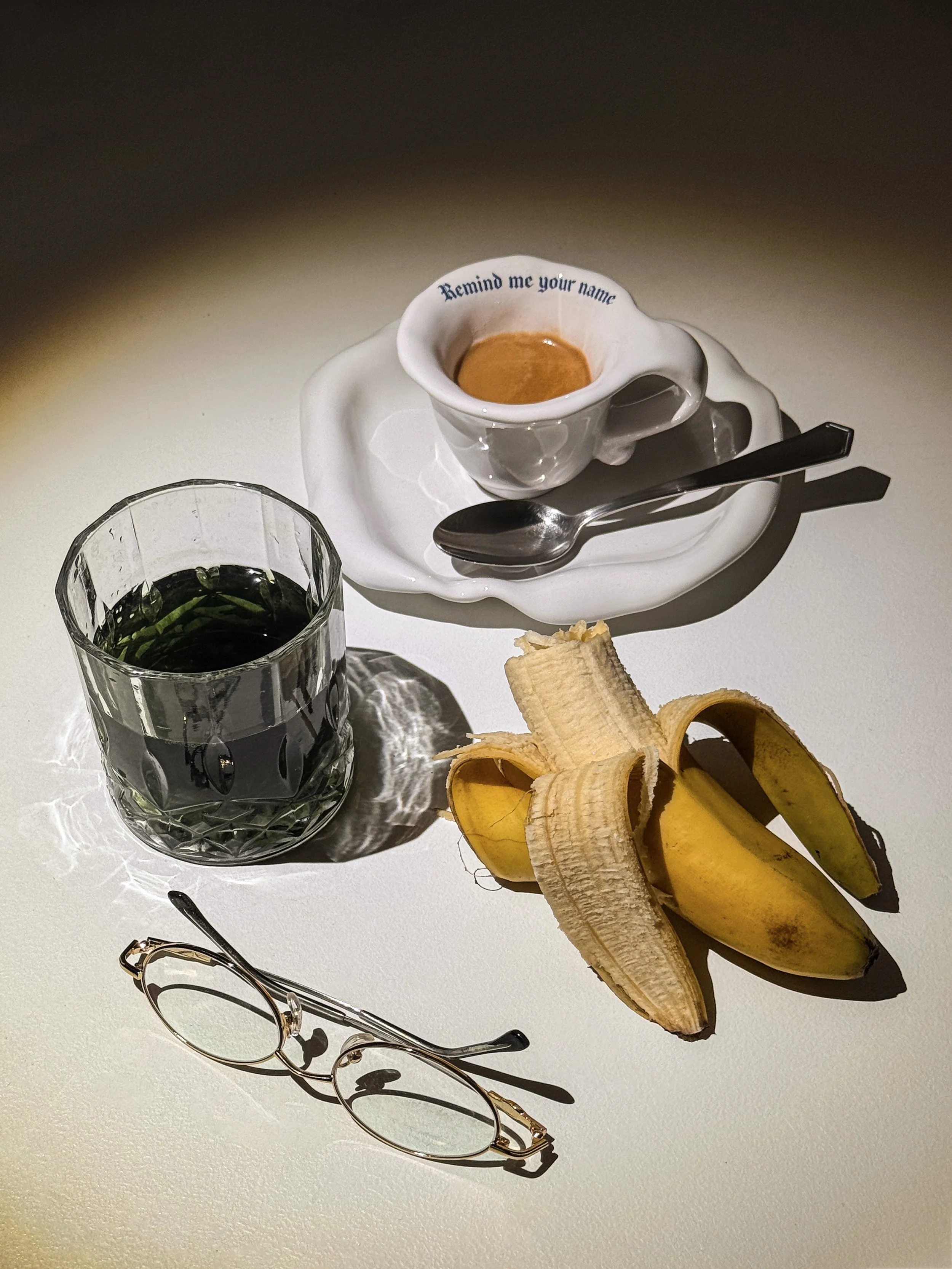 A white table holding a cup of espresso with a saucer, a spoon, a glass of green liquid, a peeled banana, and a pair of glasses.