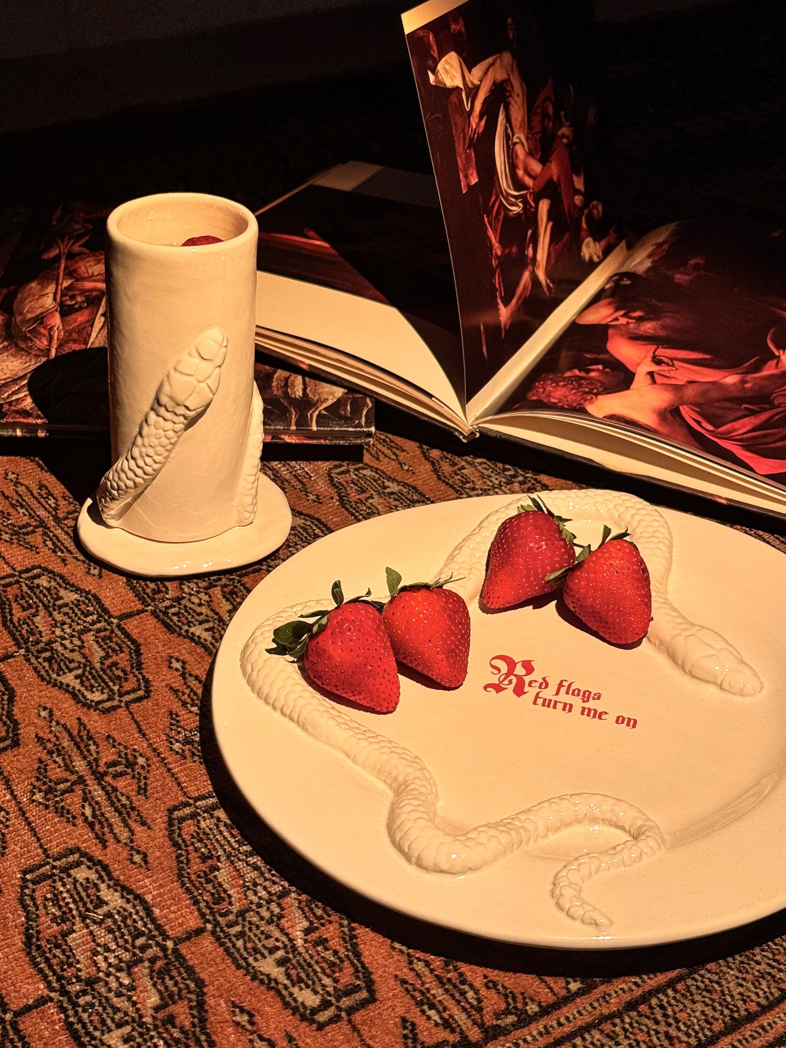 A white plate with four strawberries and a snake-shaped white icing decoration with red writing, placed on a patterned rug. Behind it, an open photo album and a cup with a red object inside, all illuminated by warm, dim lighting.
