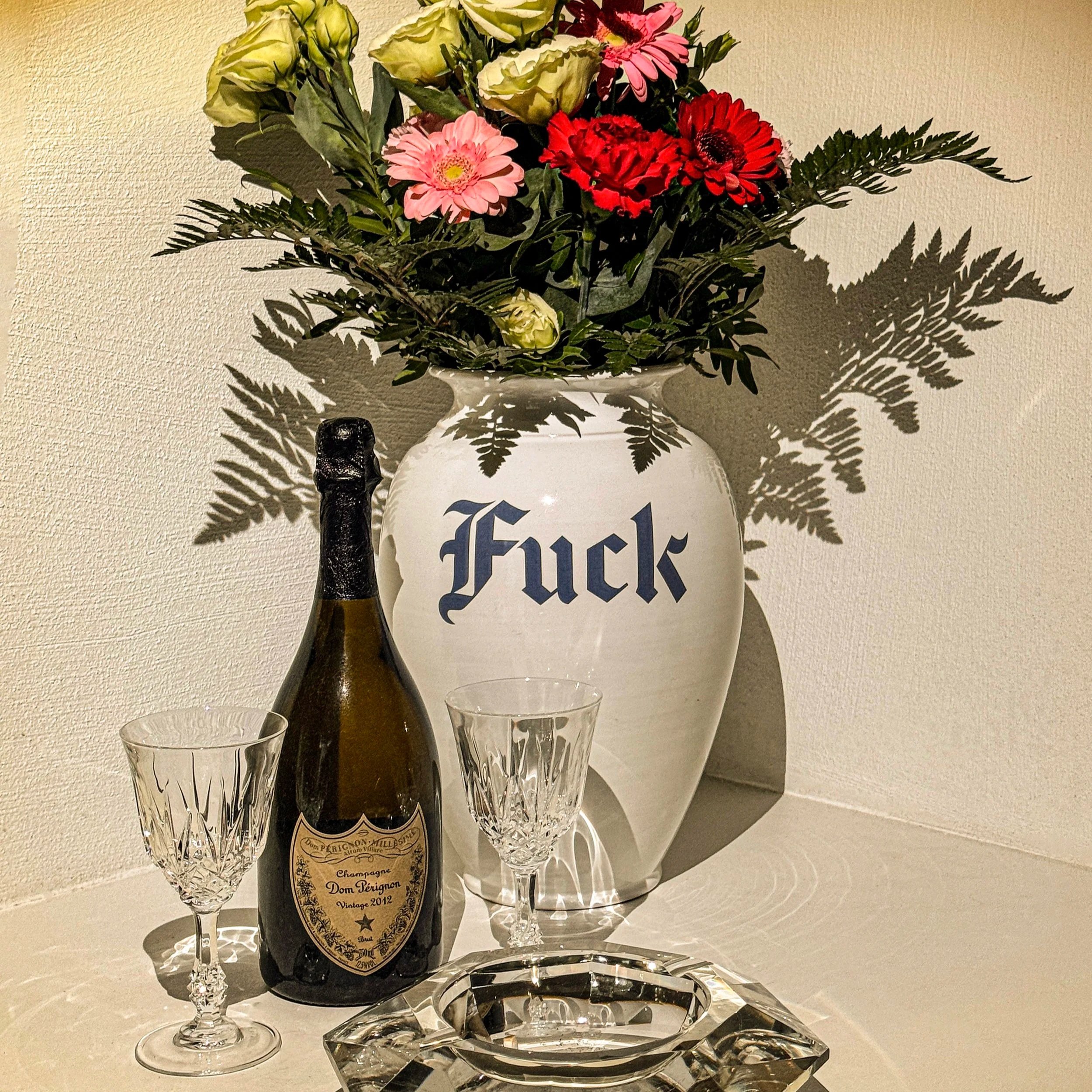 A white vase with blue lettering containing a bouquet of light green, pink, and red flowers with fern leaves, along with two wine glasses and a champagne bottle on a glass tray.