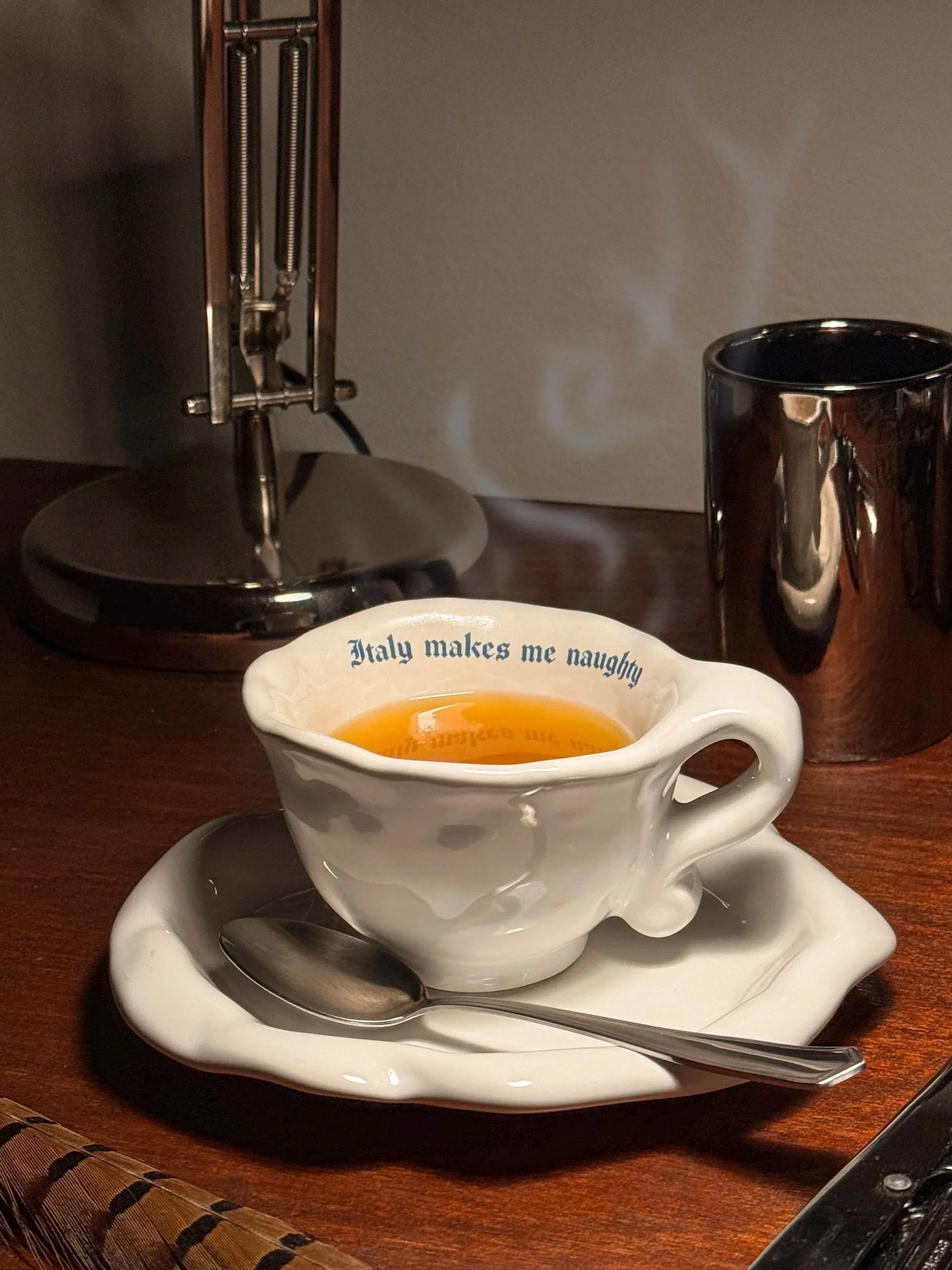 A cup of tea with the phrase "Italy makes me naughty" on the rim, placed on a matching saucer with a spoon, on a wooden table next to a metallic coffee mug and a vintage-style lamp.