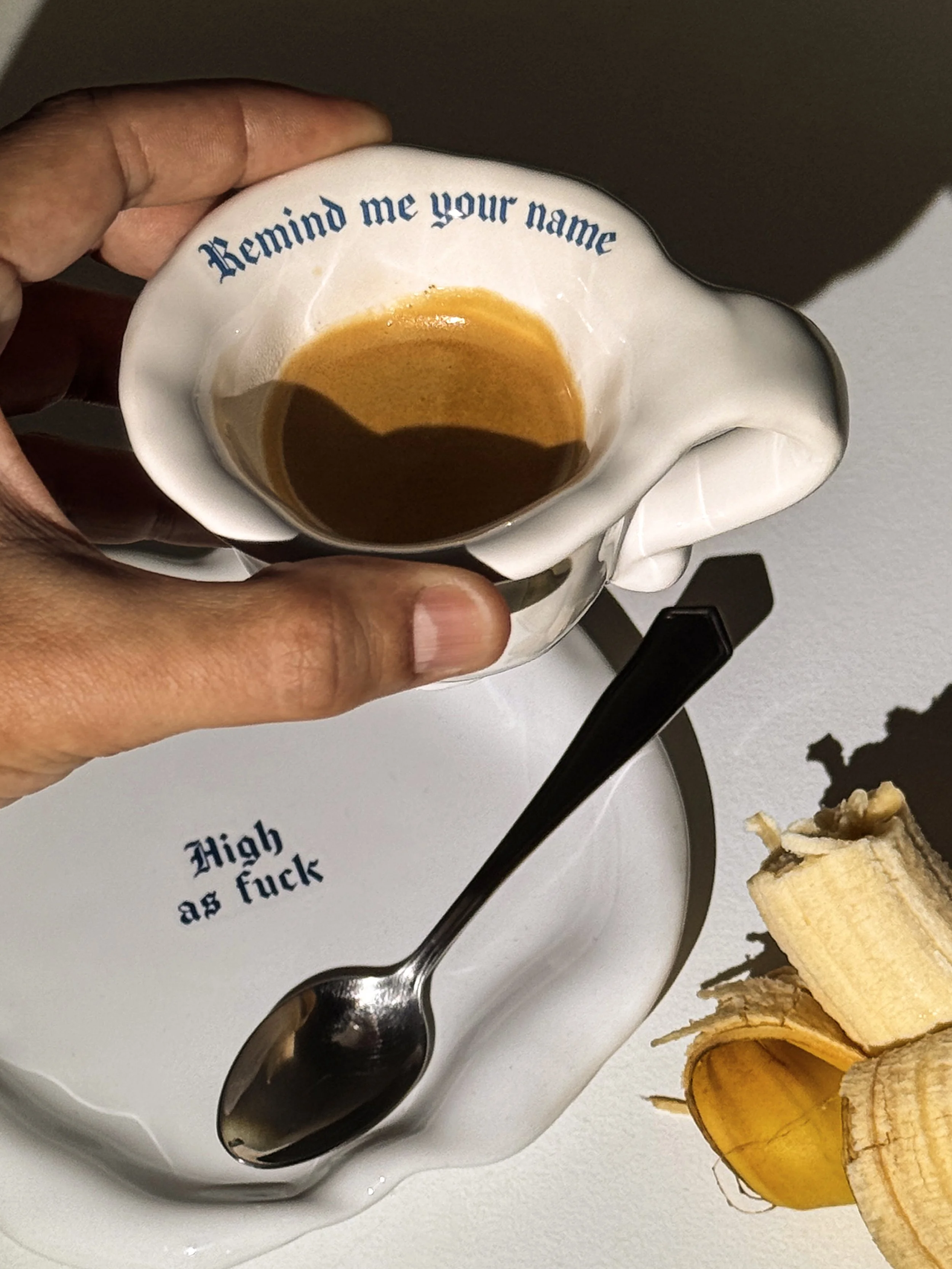 A hand holding a teacup with the phrase "Remind me your name" written on it, containing some leftover coffee, placed on a saucer with the phrase "High as fuck" and a spoon, and a banana peel on a white surface.