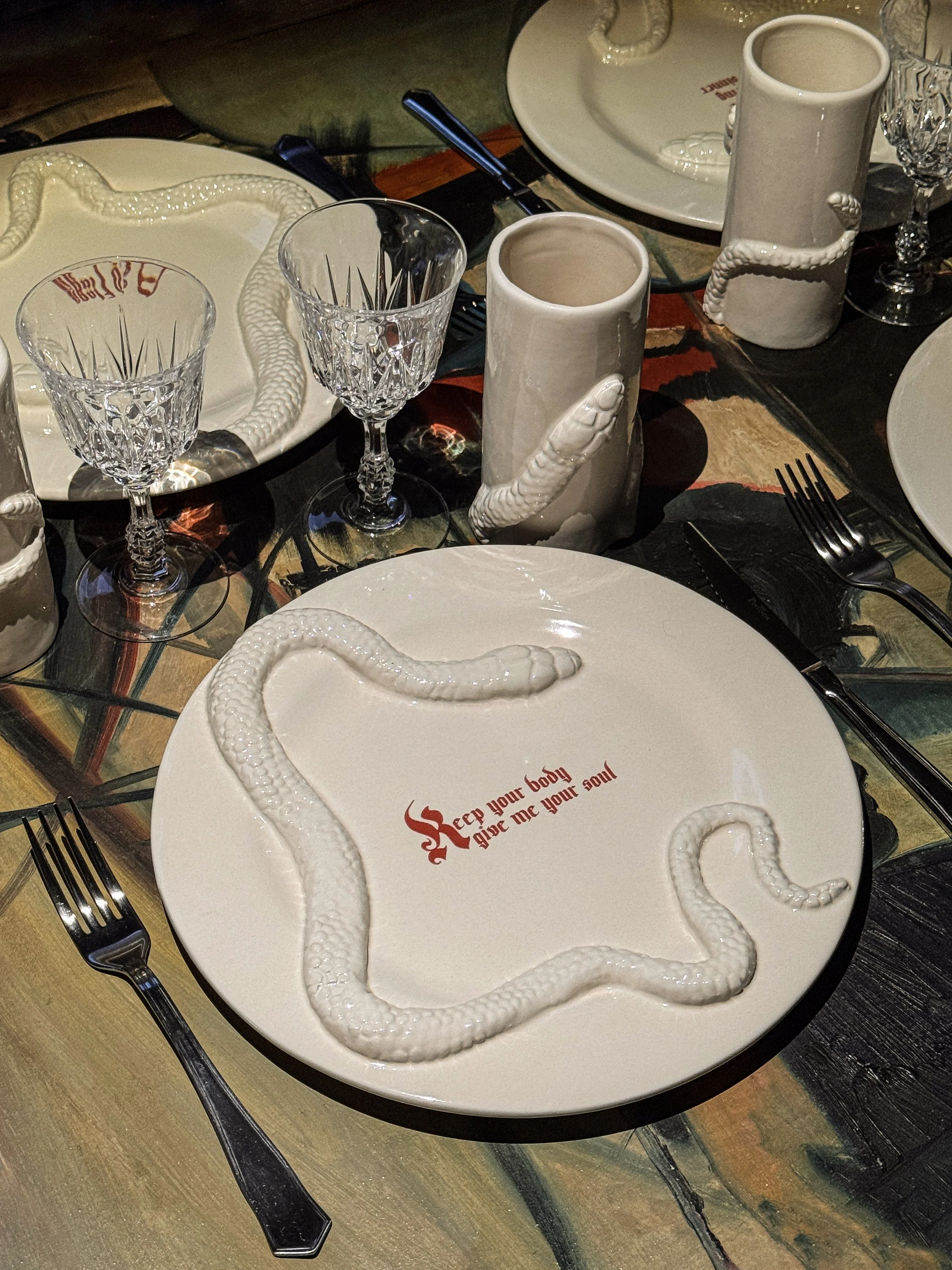 Table setting with white plates, crystal glassware, black utensils, and unique white mugs with a snake motif. The plates have the phrase 'Keep your body give me your soul' printed in red.