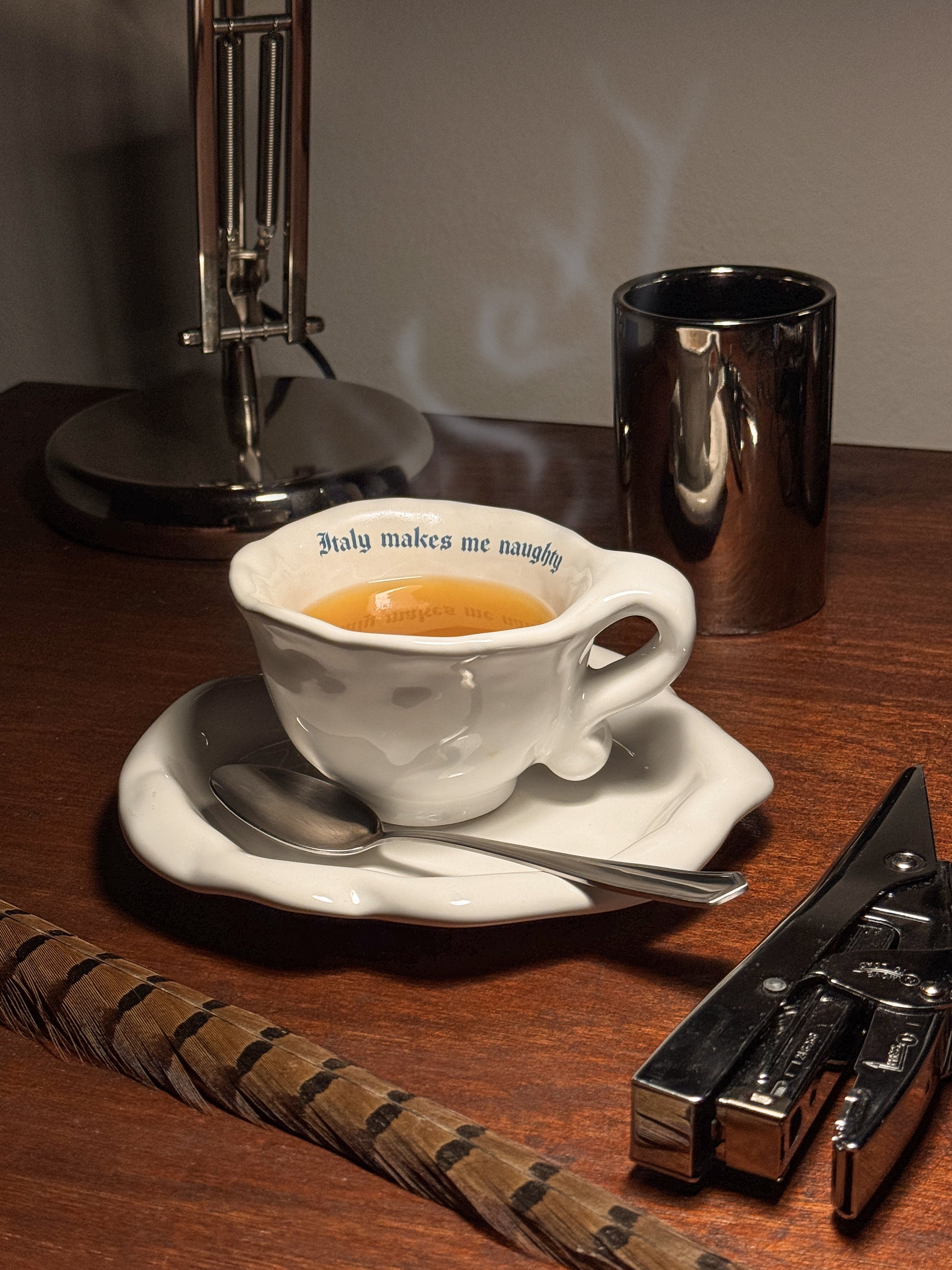 A steaming cup of tea on a fancy saucer with a spoon, with the phrase 'Italy makes me naughty' on it, on a wooden desk. Also on the desk are a black mug, a stapler, and a lamp in the background.